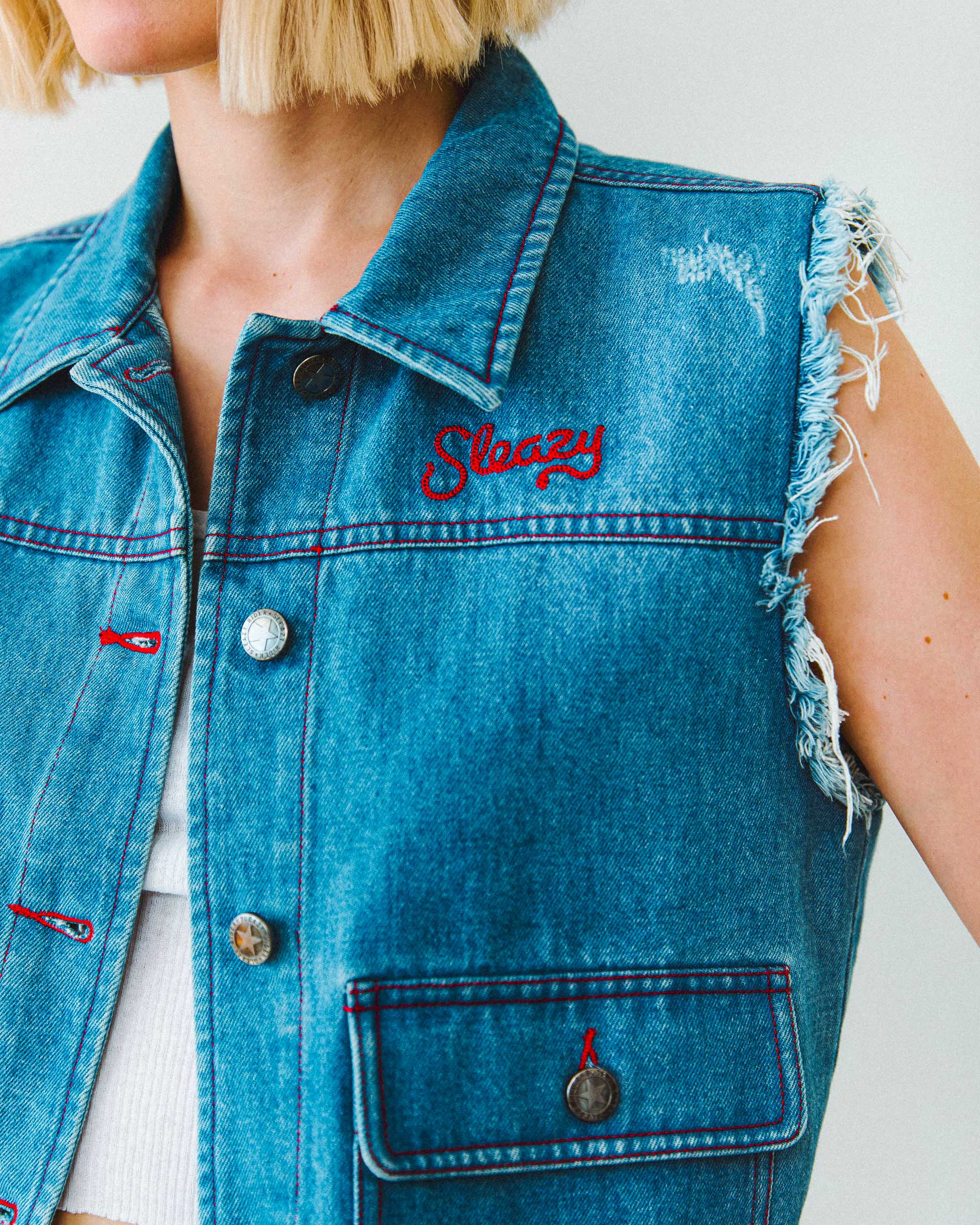 A woman wearing a denim cut off vest with red stitching and red "Sleazy" embroidery.