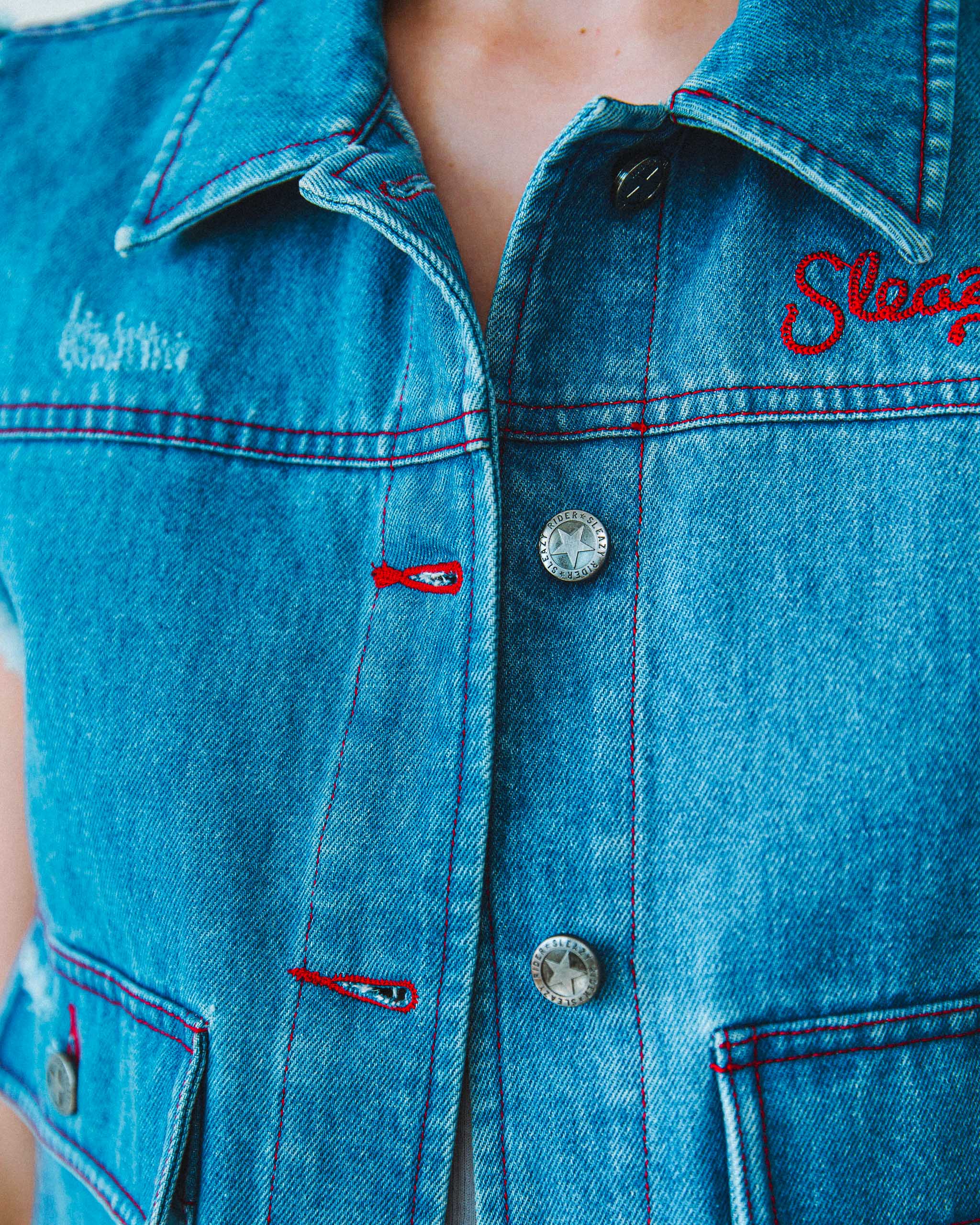 Close-up of a blue denim jacket with embroidered "Sleazy" text and star buttons.