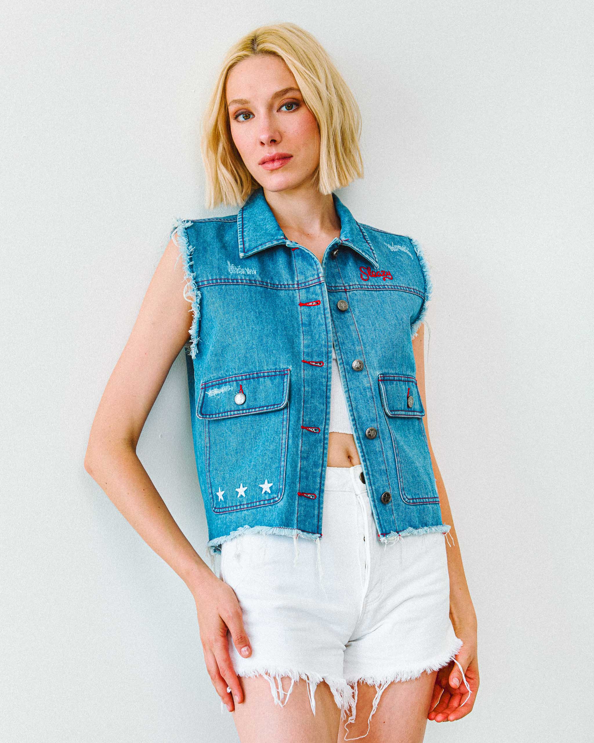 A young woman wearing a denim cut off vest with red stitching and red "Sleazy" embroidery.