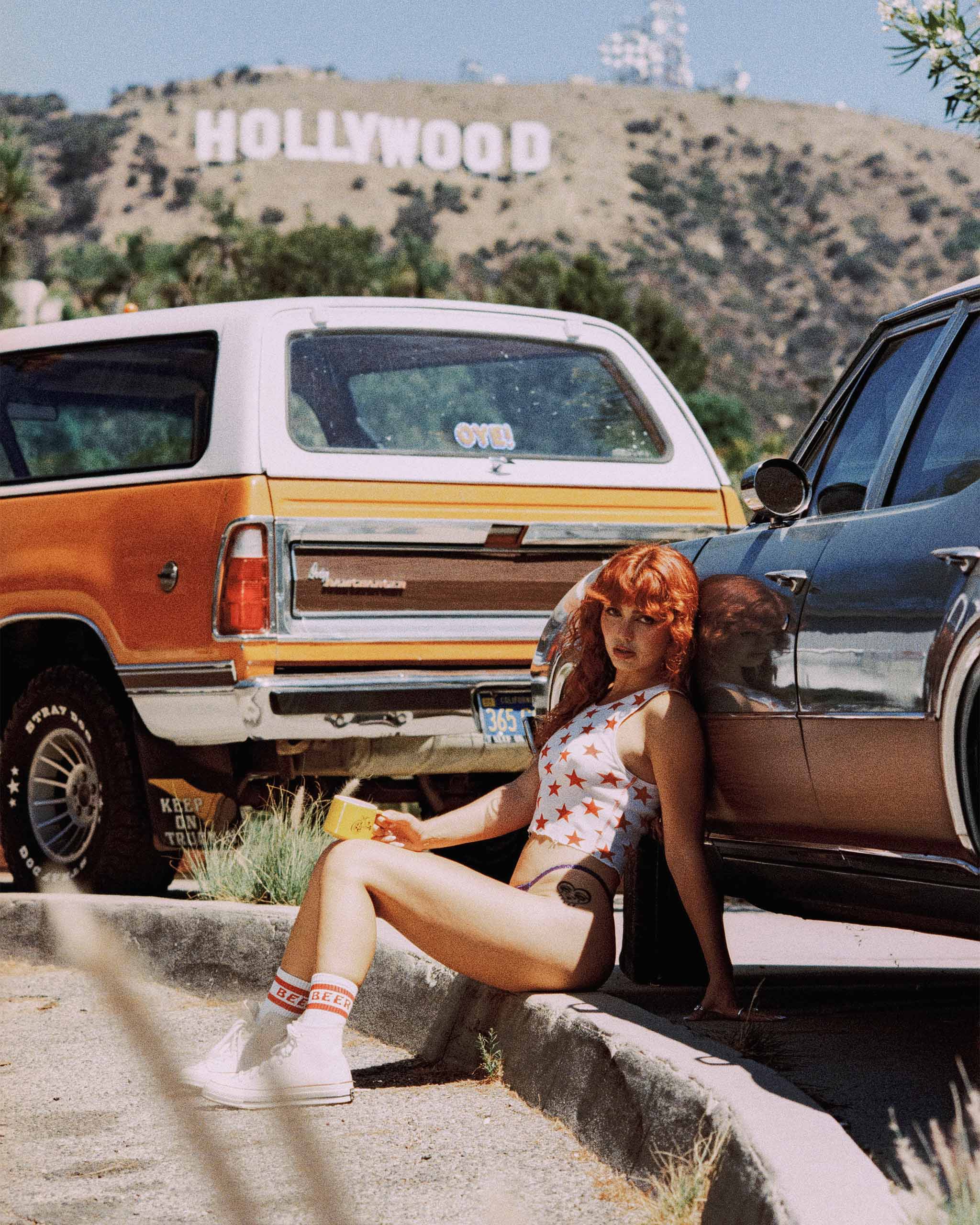 Person with red hair leaning against a vintage car with the Hollywood sign in the background wearing Sleazy Rider "Beer" socks and Starstudded tank top.