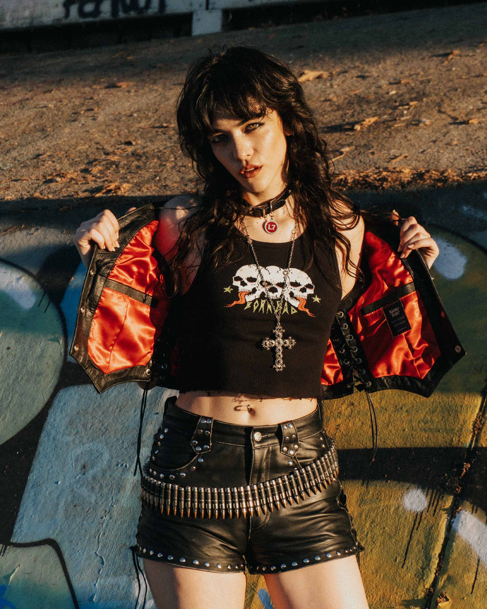 Person wearing a black crop top with skull design with a matching studded leather set, against a graffiti-covered wall.