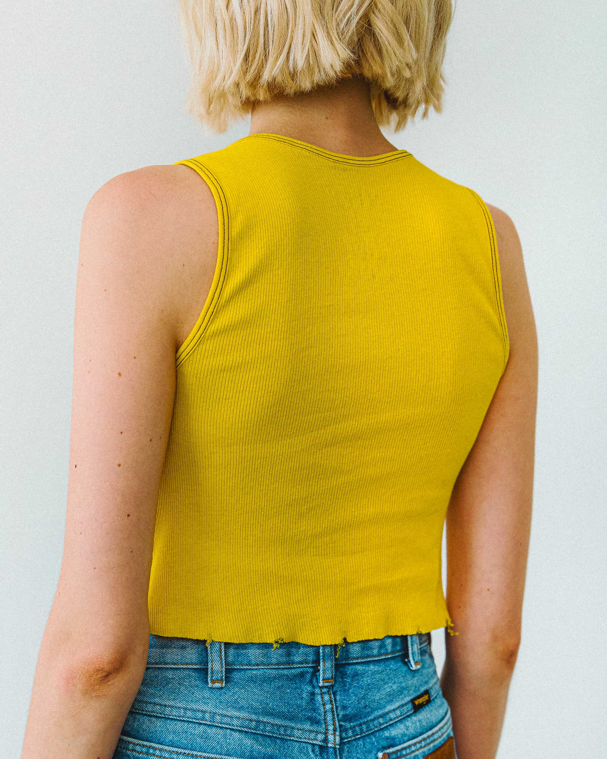 Person wearing a yellow sleeveless tank top and blue jeans against a plain background