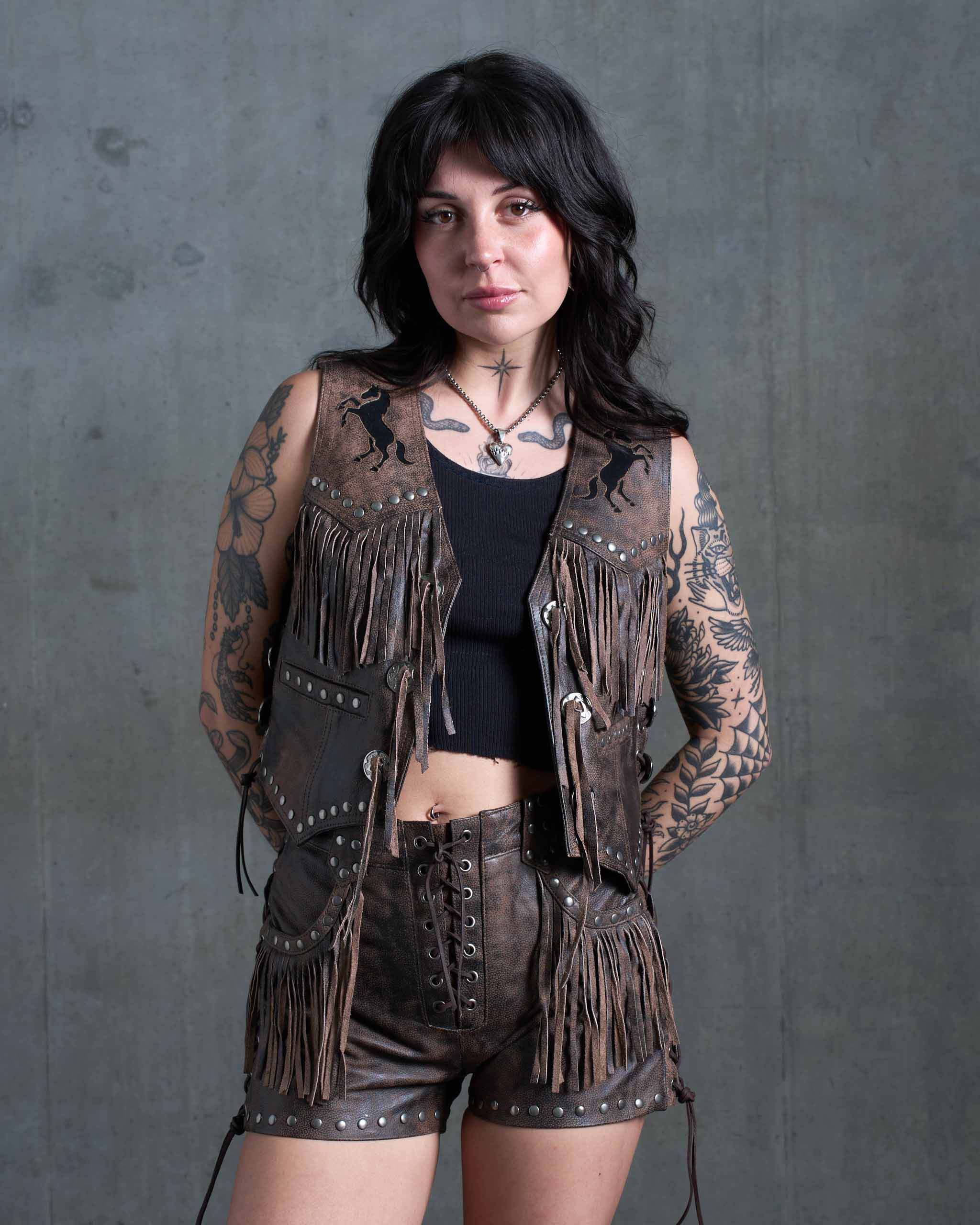 A tattooed model wearing the Sleazy Rider Colt Studded Fringe Leather vest and shorts set with a concrete backdrop.
