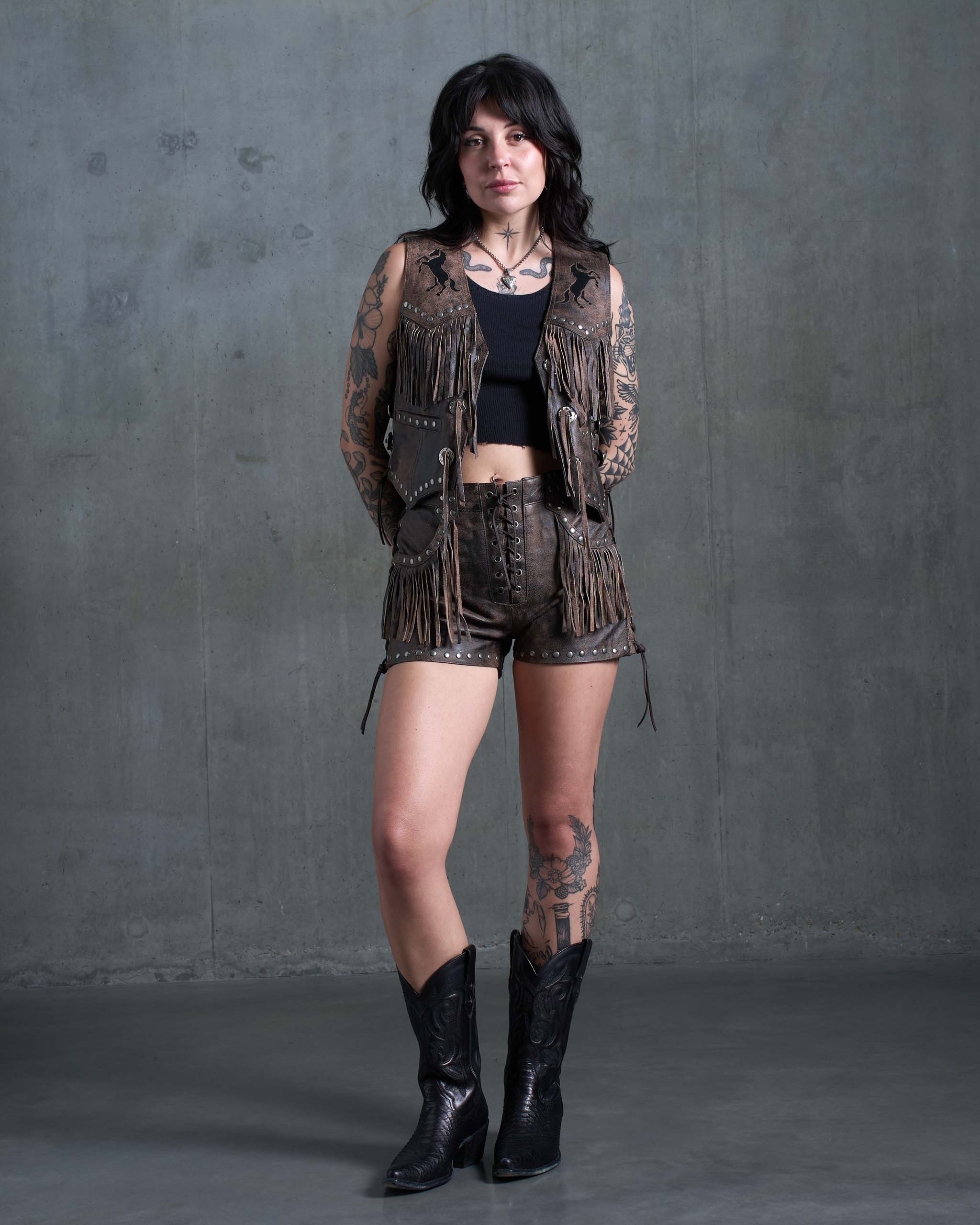 A tattooed model wearing the Sleazy Rider Colt Studded Fringe Leather vest and shorts set with a concrete backdrop.