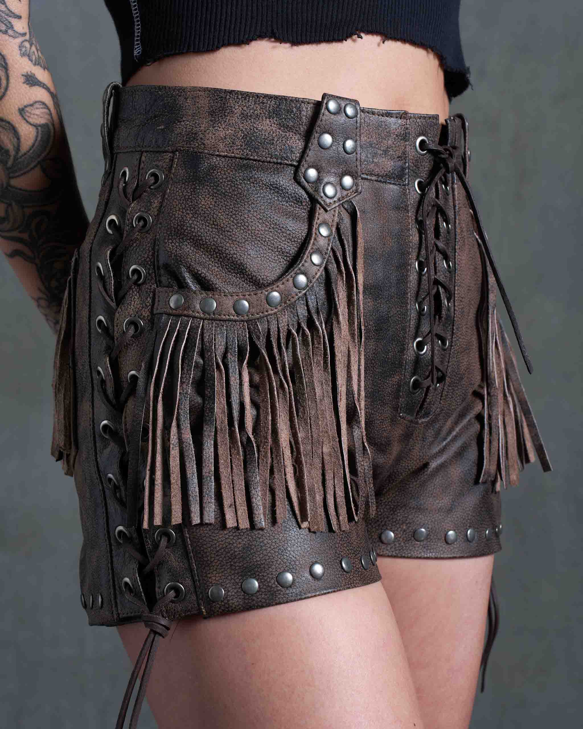 A tattooed model wearing the Sleazy Rider Colt Studded Fringe Leather vest and shorts set with a concrete backdrop.