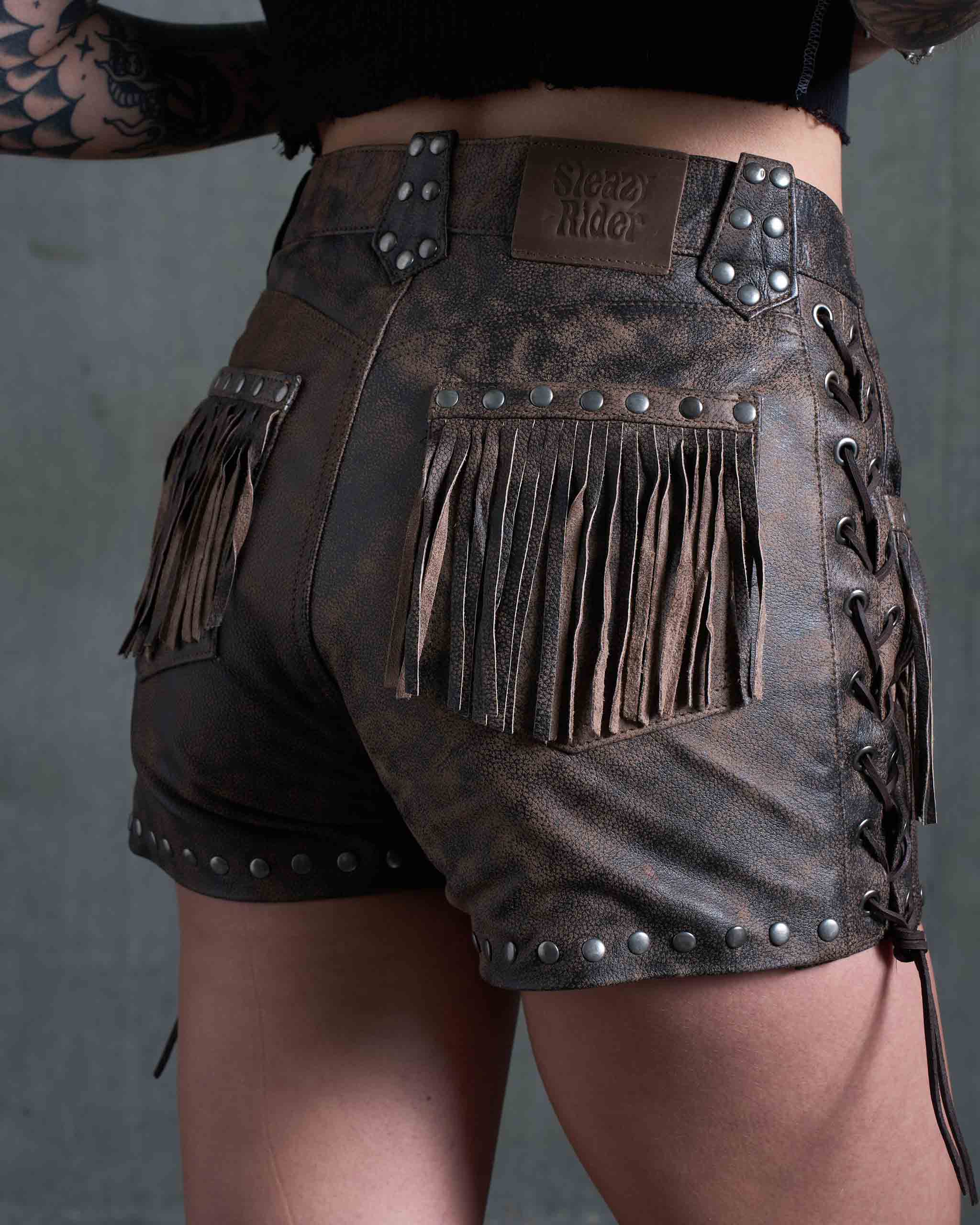 A tattooed model wearing the Sleazy Rider Colt Studded Fringe Leather vest and shorts set with a concrete backdrop.