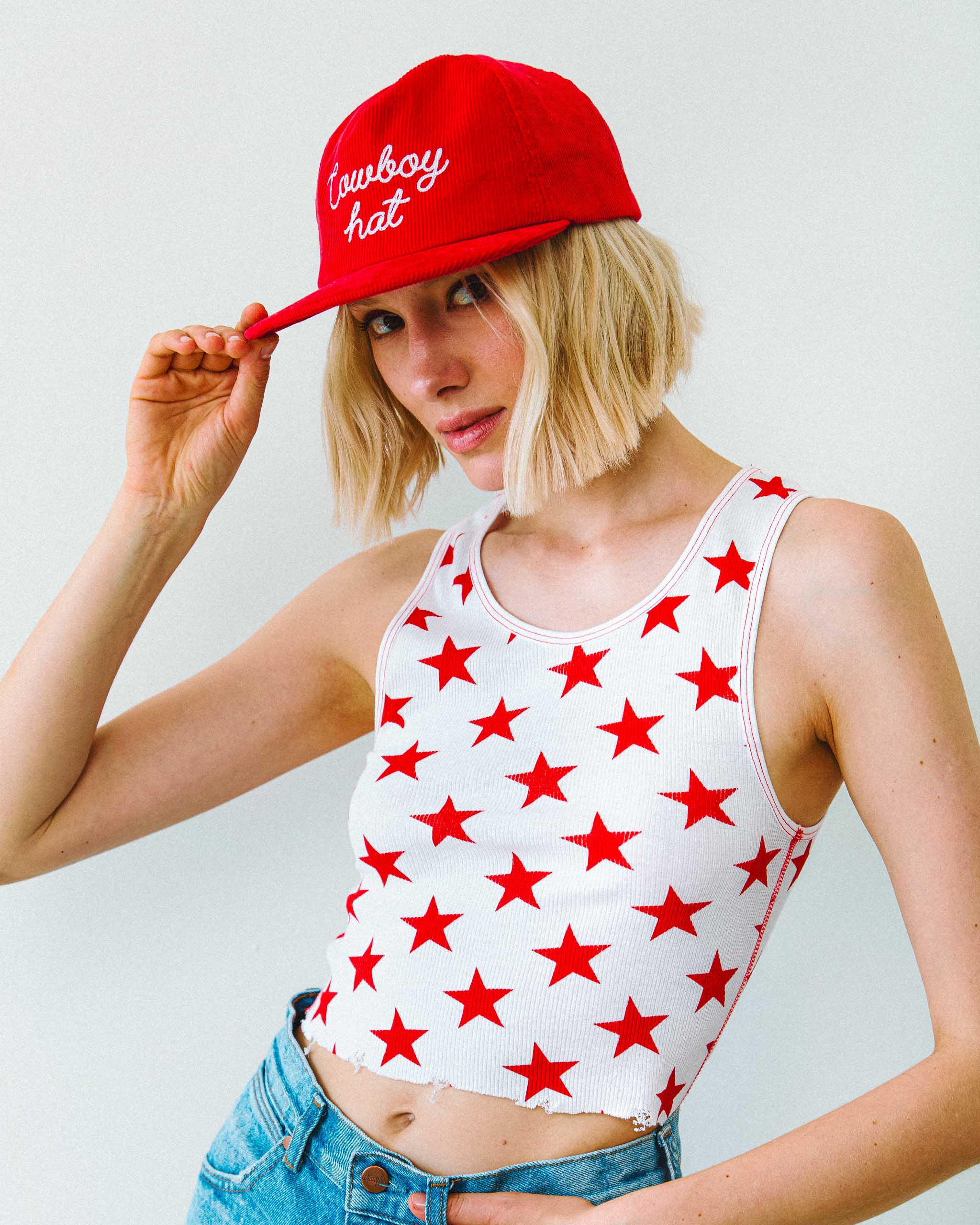A woman wearing a white tank top with red stars printed all over and a red cap with "Cowboy hat" embroidery.