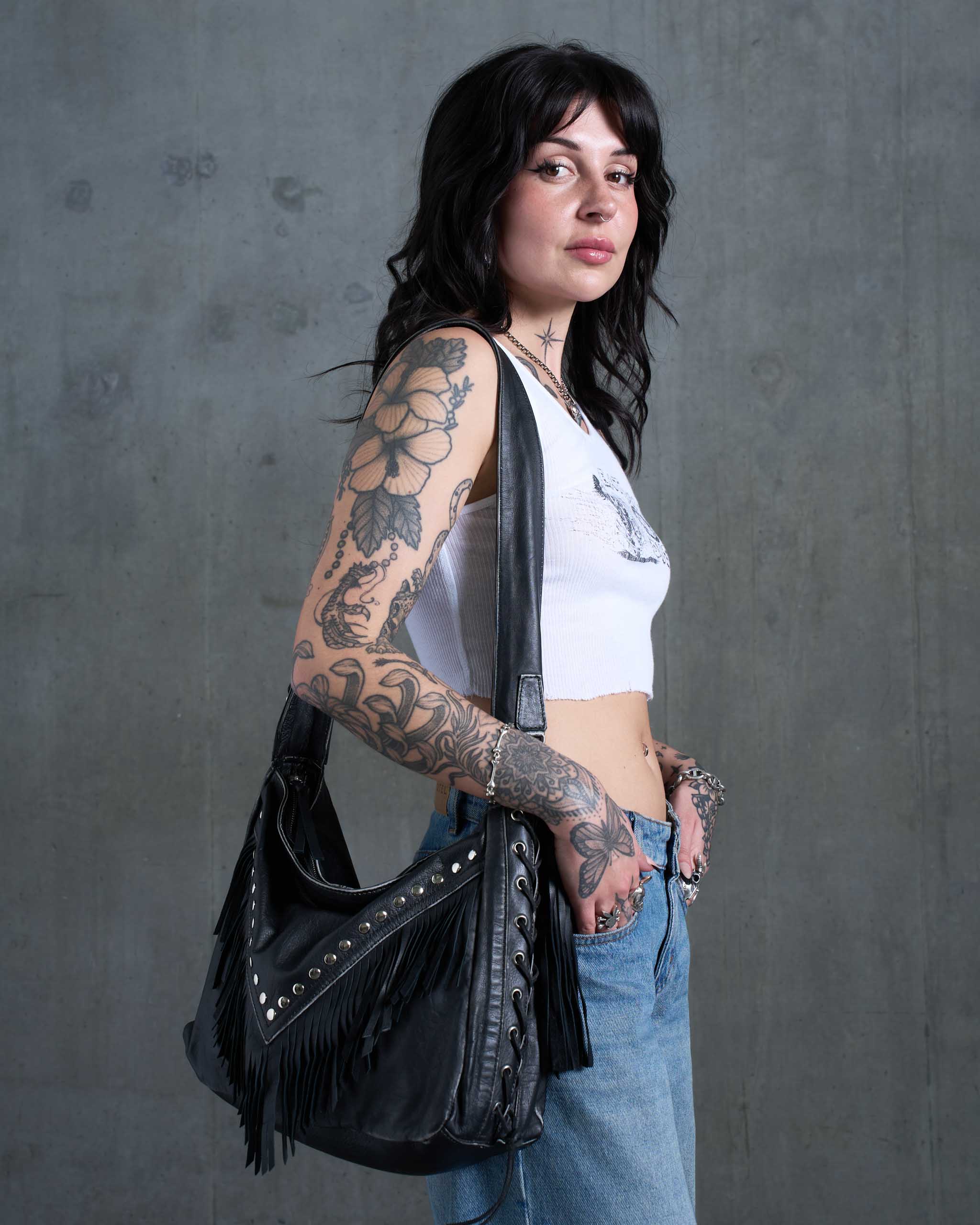 An alt model wearing a dark western slouchy bag, with studs, fringe and lace-up sides.