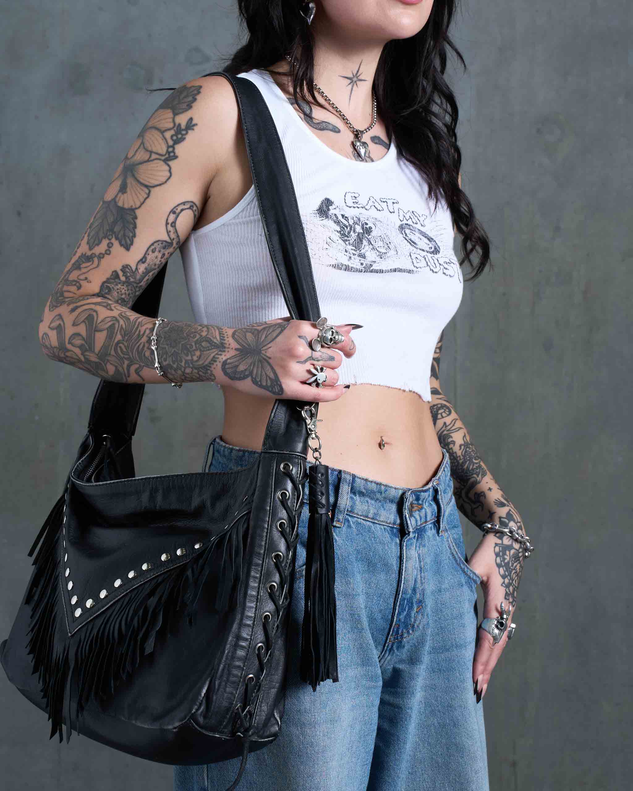 A tattooed person holding a gothic western tote bag, with studs and fringe.