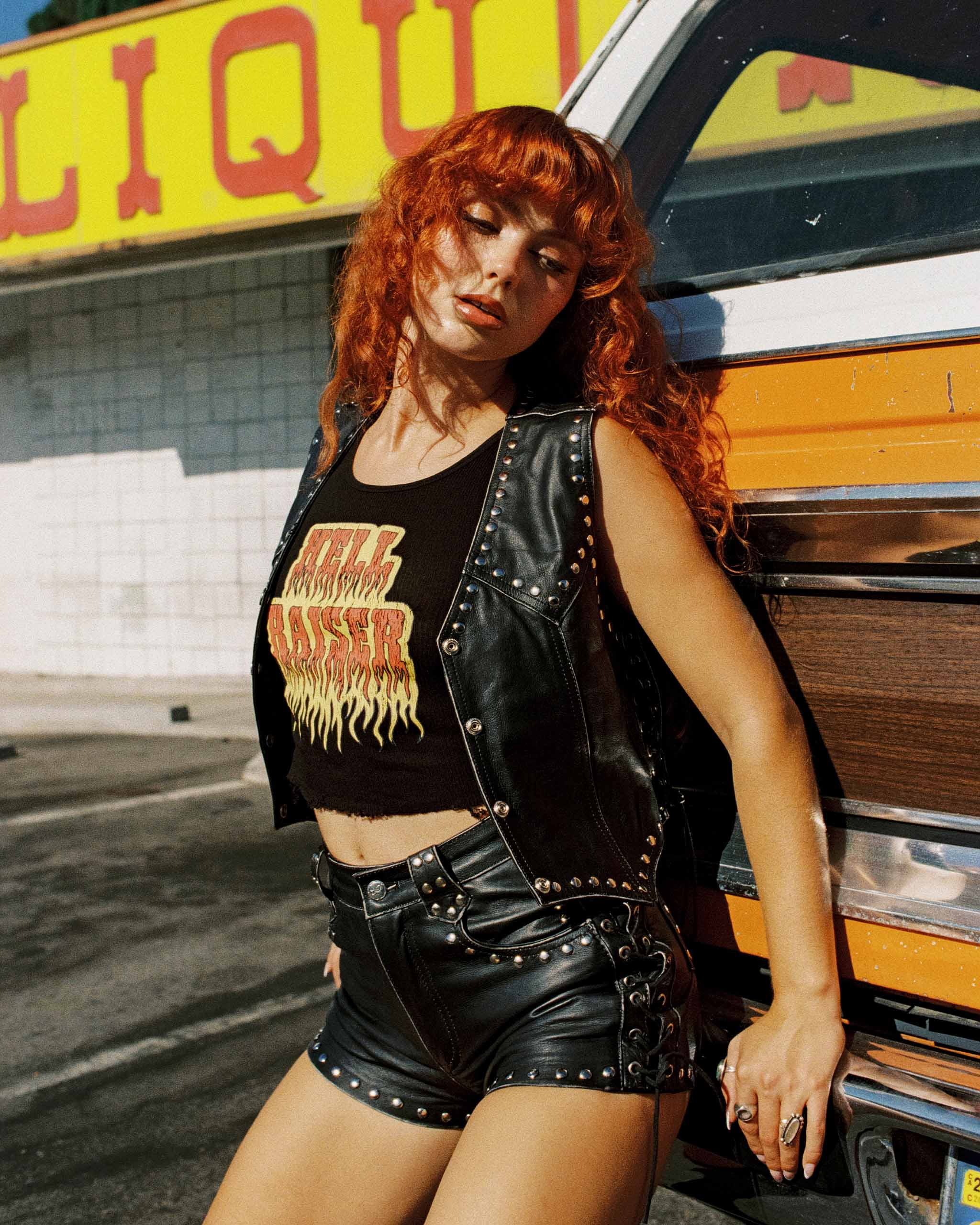 Redhead model wearing a black leather studded vest and shorts standing next to a vintage orange truck with 'Liquor' sign in the background.