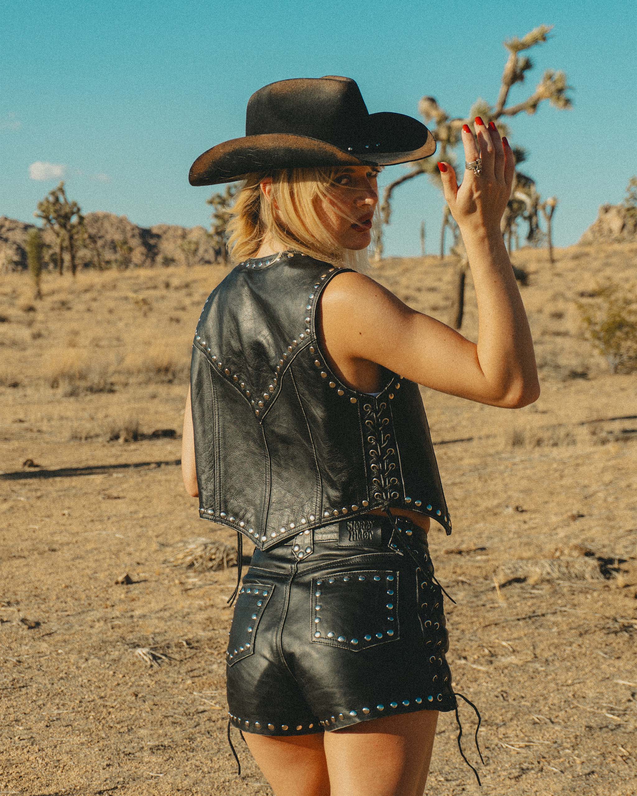 A cowgirl wearing the Sleazy Rider coyote studded leather vest and shorts in the desert with a gothic western cowboy hat.