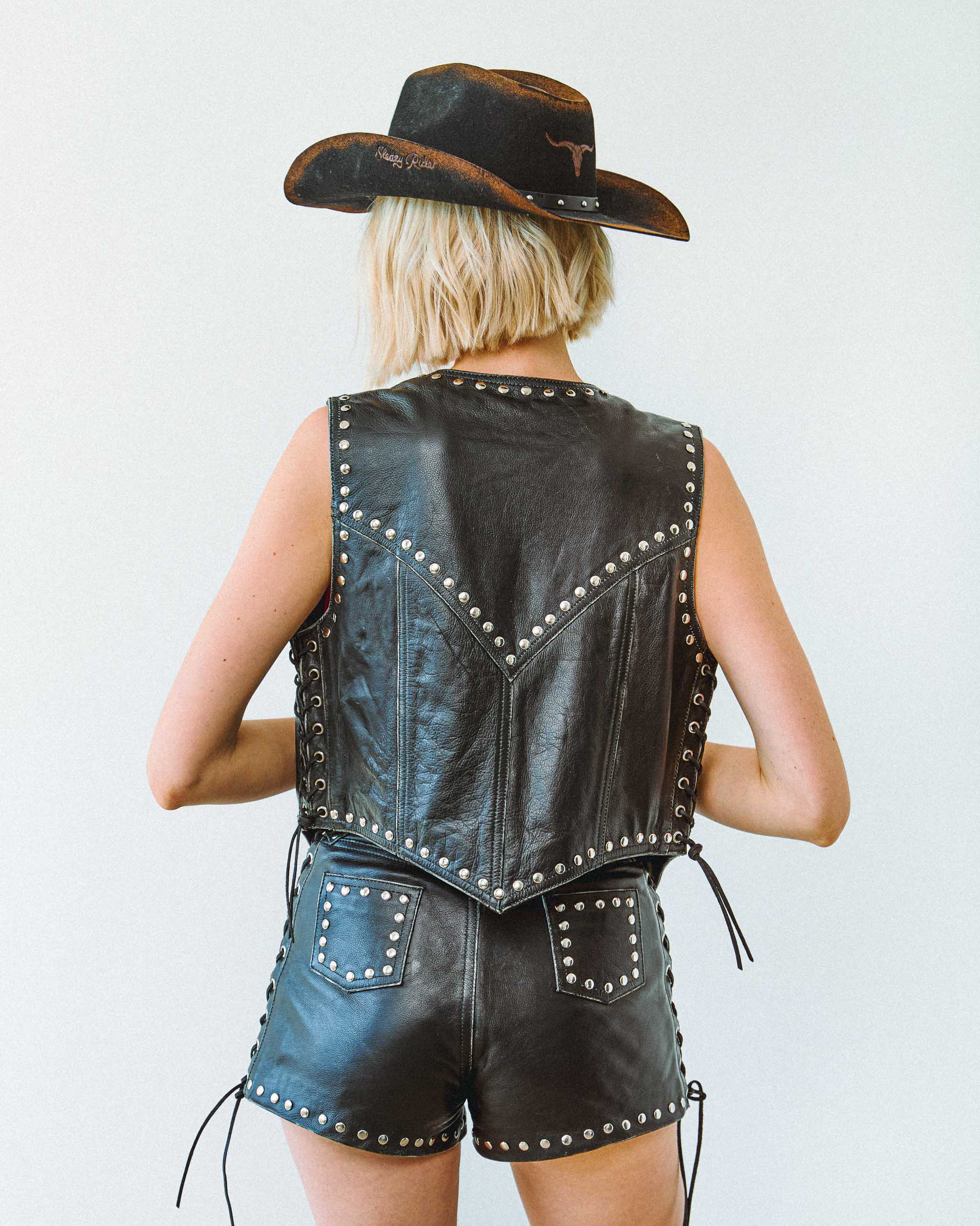 Person wearing a black studded leather vest and shorts with a cowboy hat on a white background.