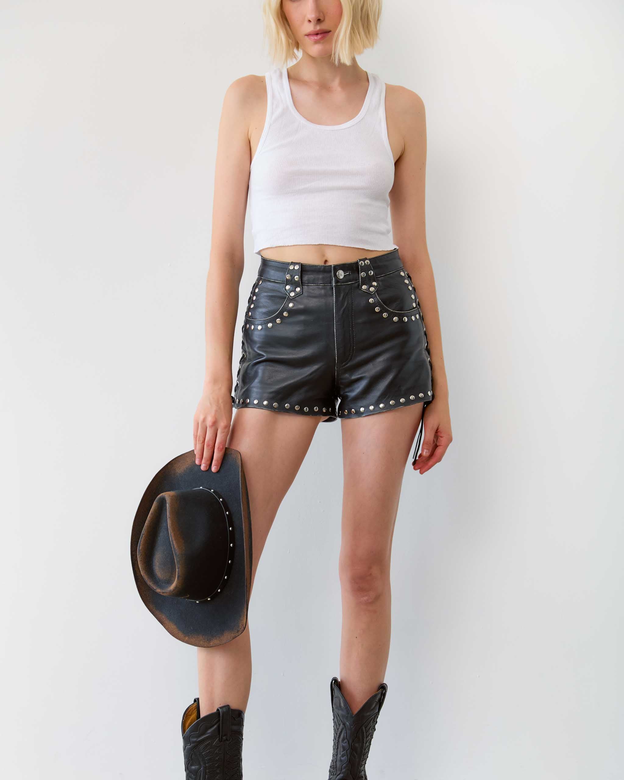 Person wearing black leather shorts with studs, a white tank top, and cowboy boots, holding a black cowboy hat.