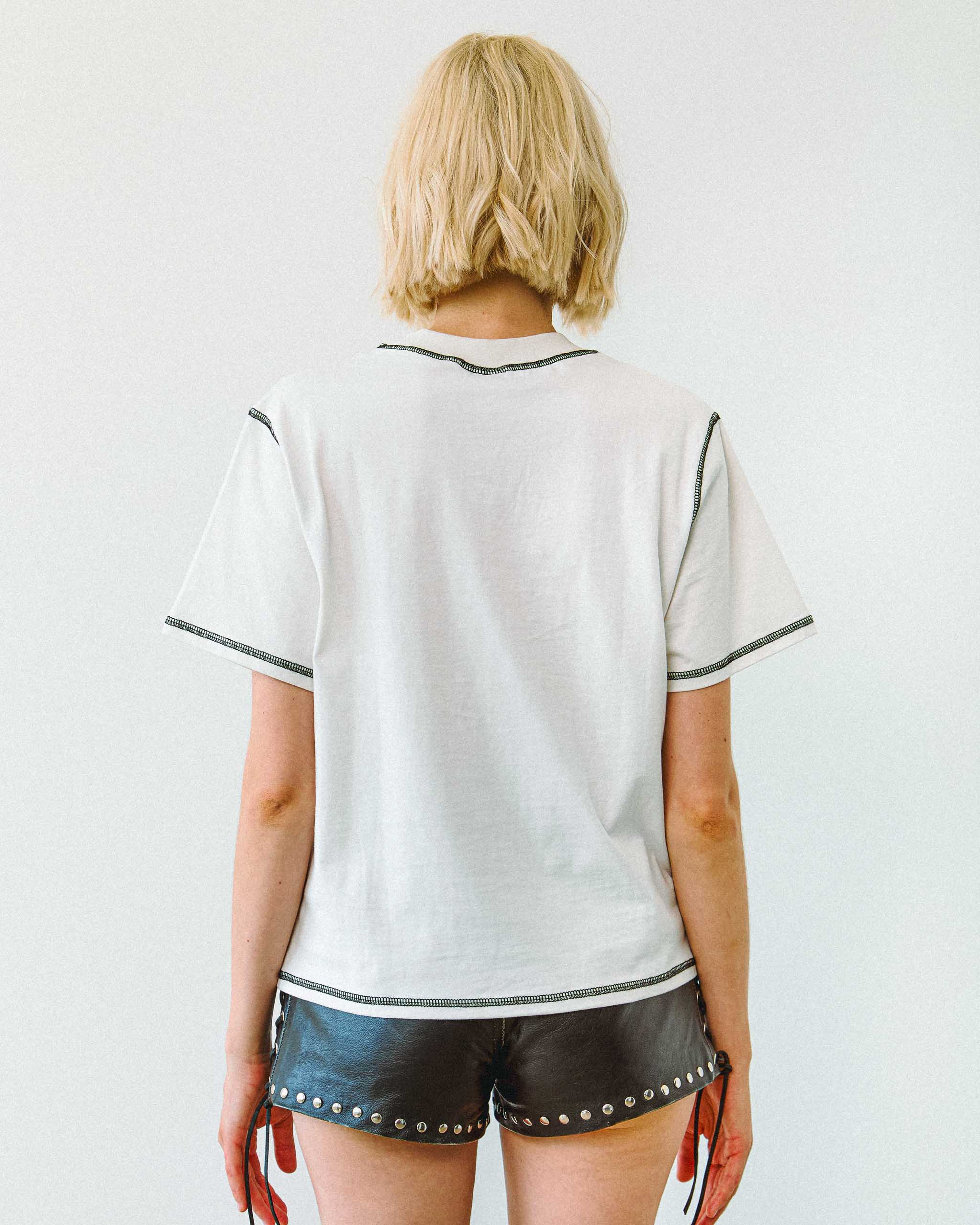 Person wearing a white shirt with black trim and Sleazy Rider black studded leather shorts with studs on a plain background.