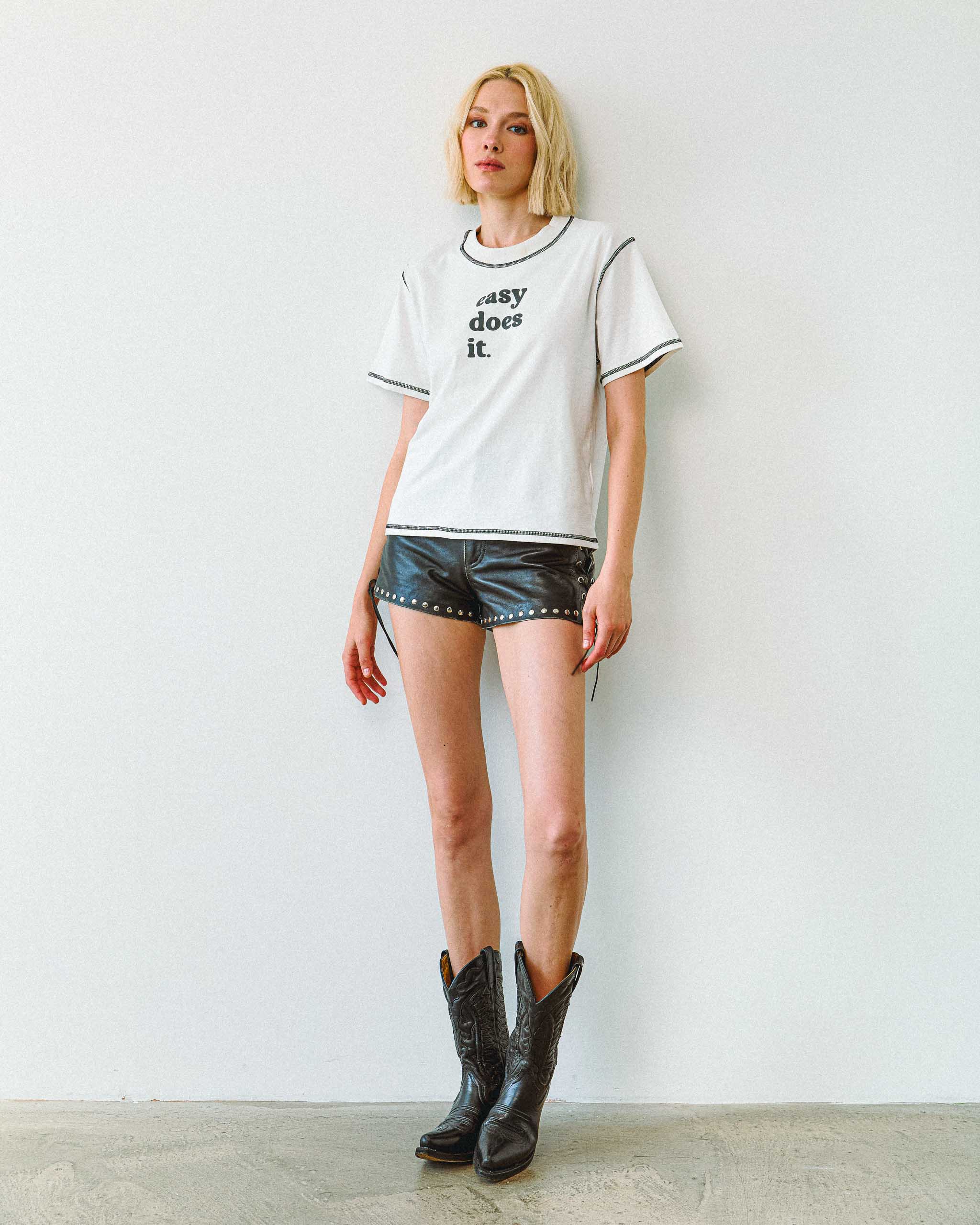An alt woman wearing a white t-shirt with 'easy does it' text, Sleazy Rider studded leather shorts and cowboy boots.