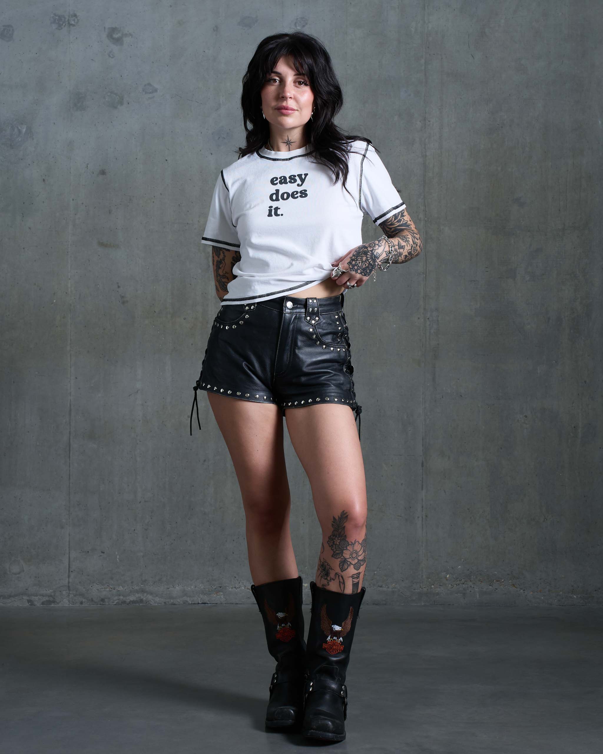 A tattooed woman wearing an 'easy does it' vintage t-shirt, studded leather shorts and black cowboy boots.
