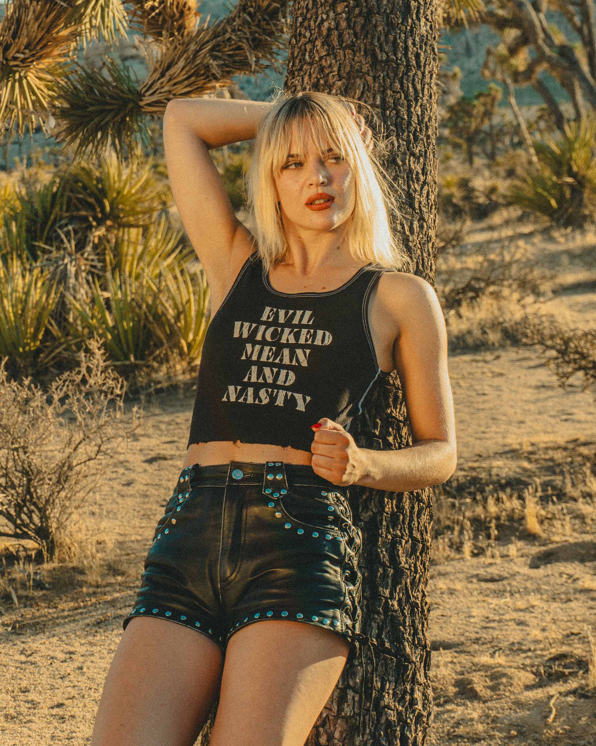Woman in the California desert leaning against a tree wearing a tank top with "Evil Wicked Mean and Nasty" text and black studded leather shorts.