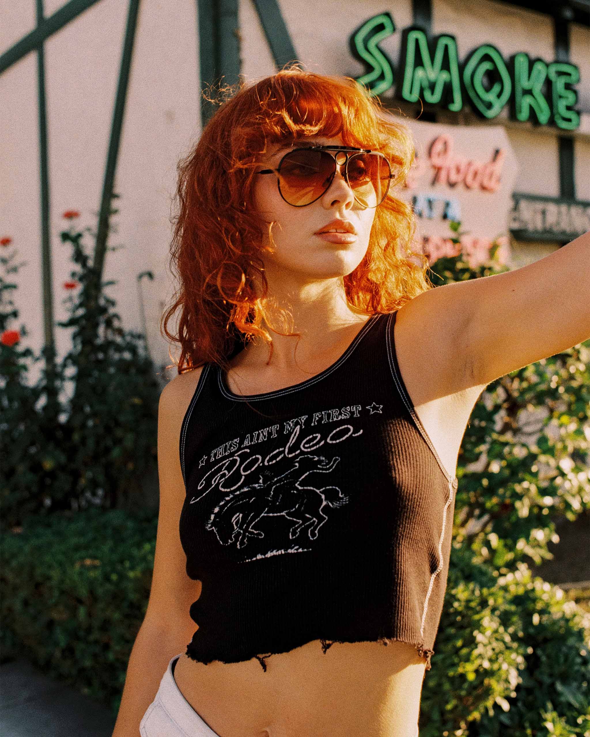 Woman with red hair wearing 70s sunglasses and a black tank top with "This Ain't My First Rodeo" text and horse graphic, standing in front of a neon sign.