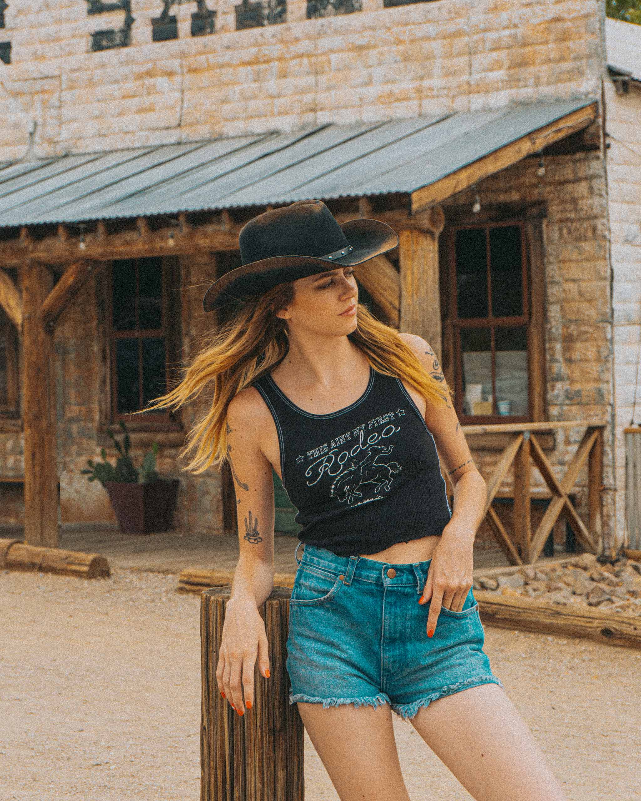 Woman in a gothic western cowboy hat and black "This Ain't My First Rodeo" tank top standing in front of an old saloon in Nevada.