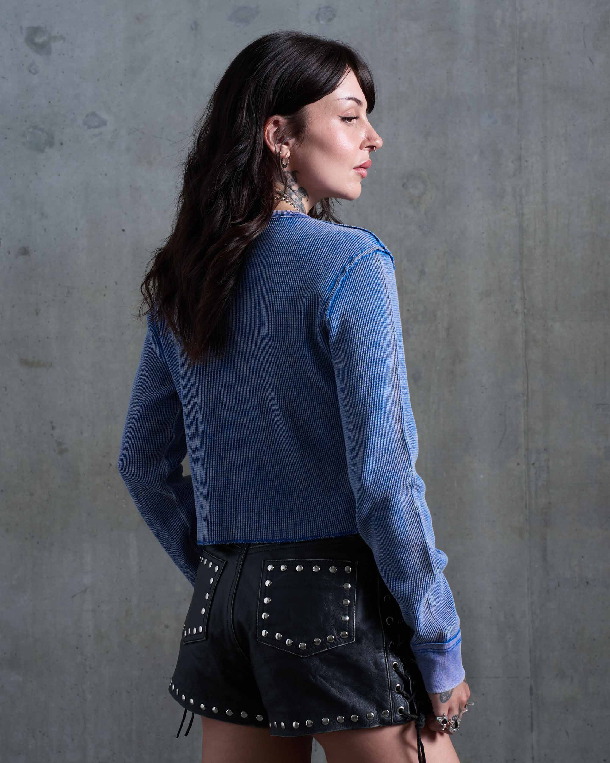 A tattooed model wearing a blue faded vintage waffle knit top, with black studded leather shorts, viewed from behind.