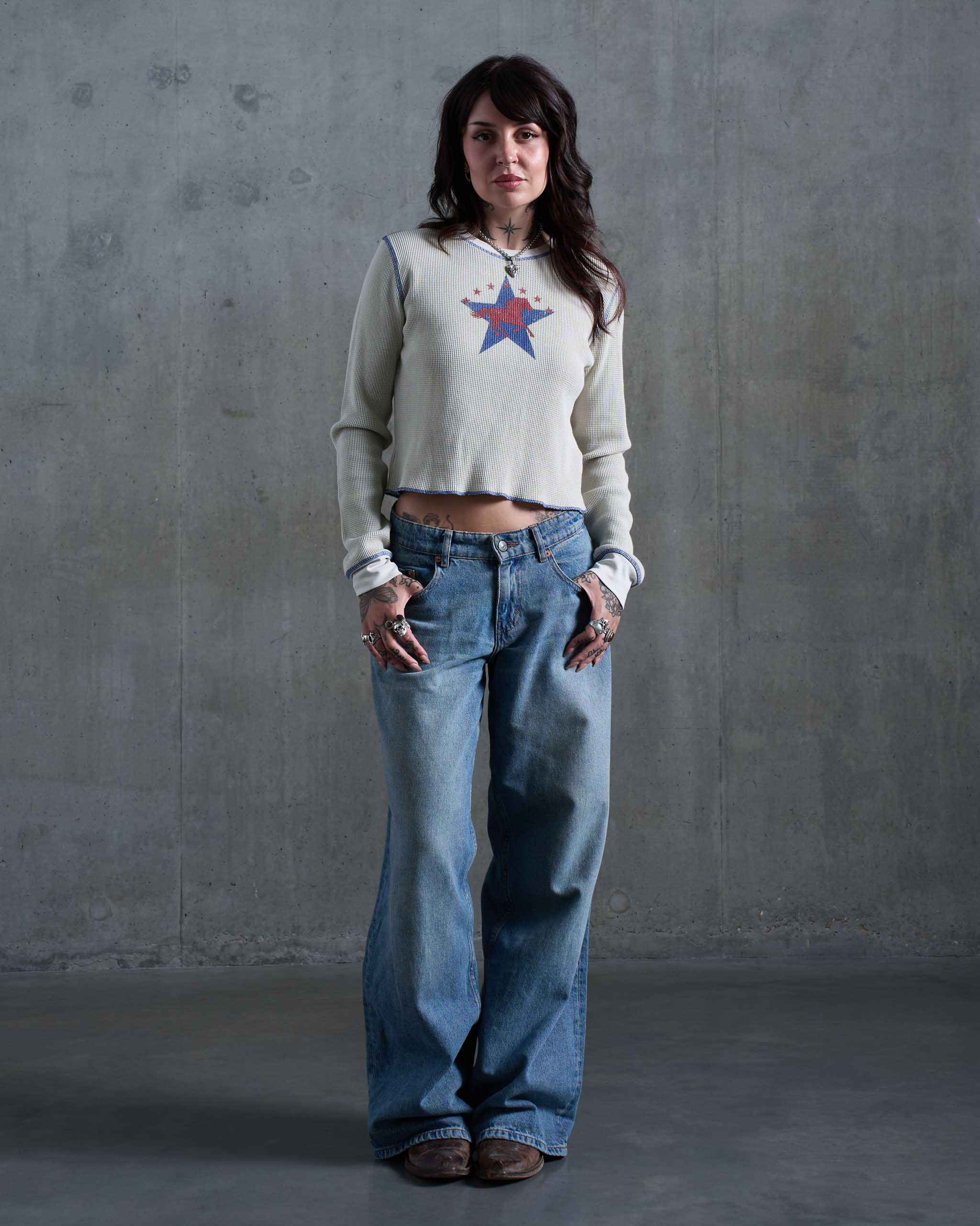 A tattooed woman wearing an off-white waffle knit top with a horse and stars print, with low rise jeans and brown cowboy boots.