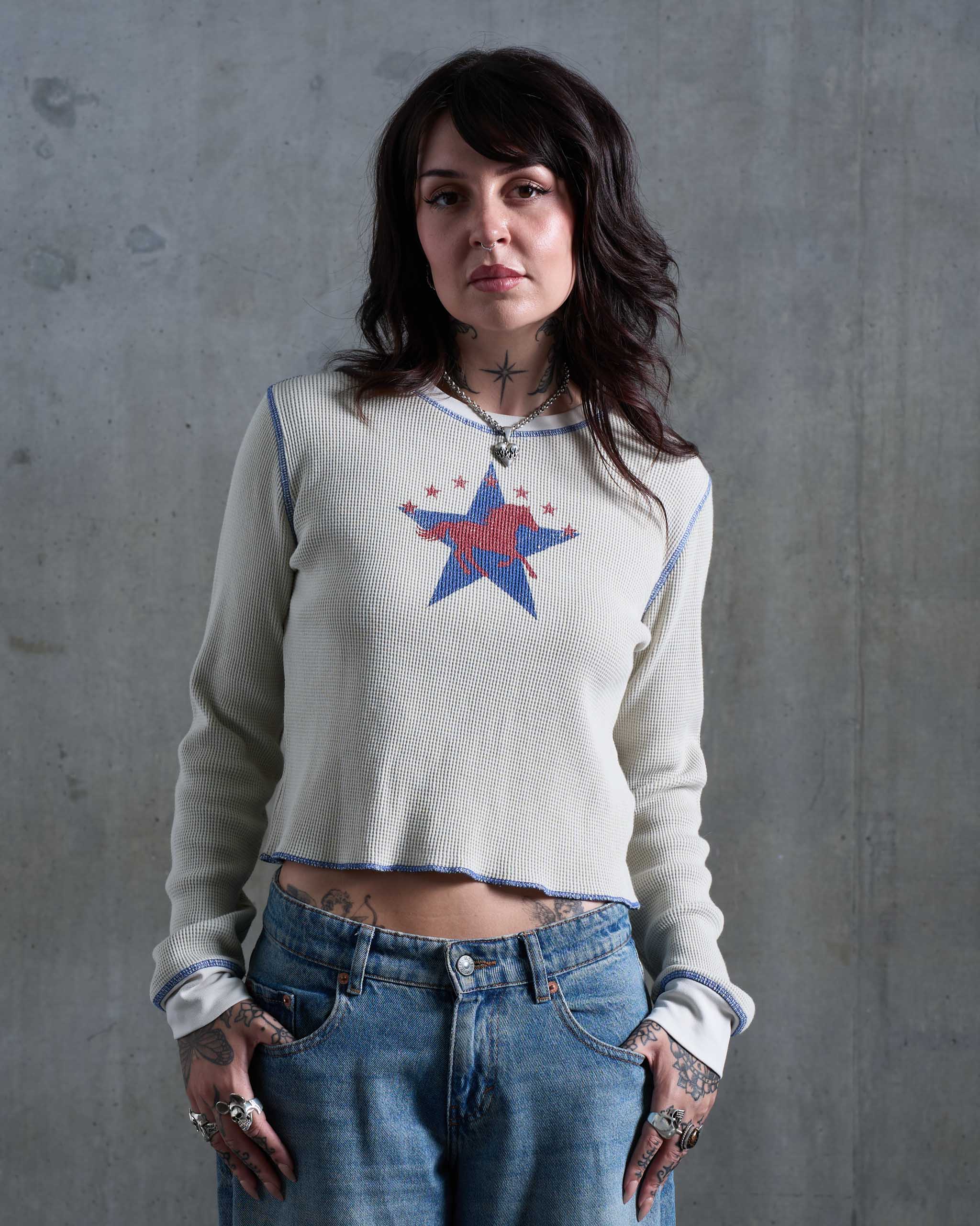 A tattooed woman wearing an off-white waffle knit top with a horse and stars print, with low rise jeans.