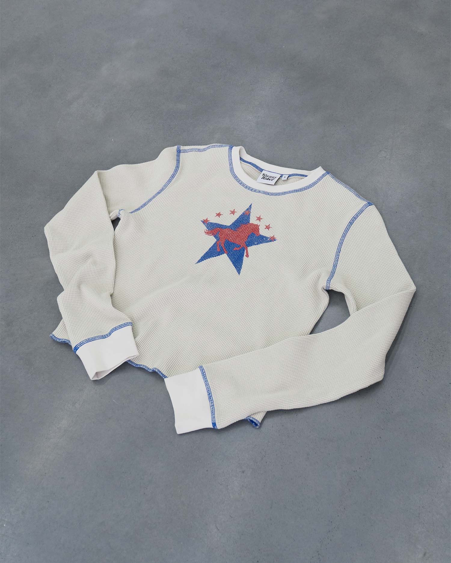 An off-white waffle sleeve top with a western horse screen print and blue contrast stitching, laid on a concrete floor.