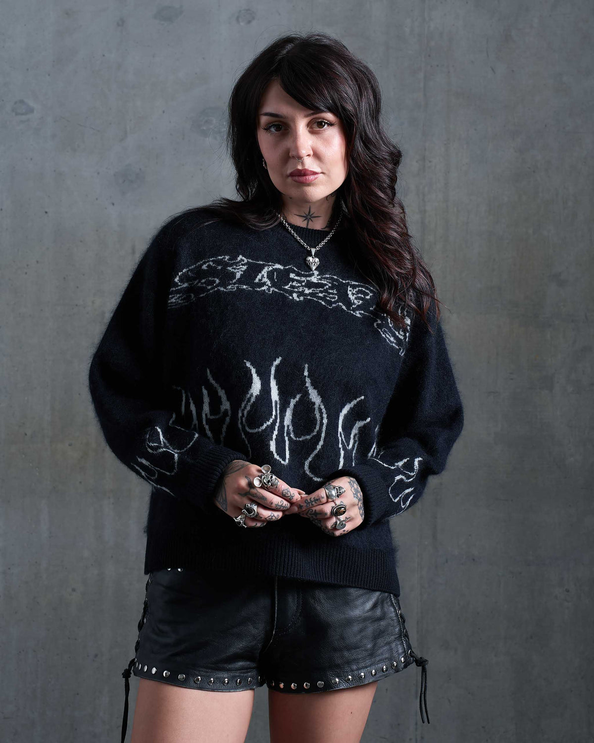 An alt woman with tattoos wearing a gothic black knitted jumper with white flames, and studded leather shorts.