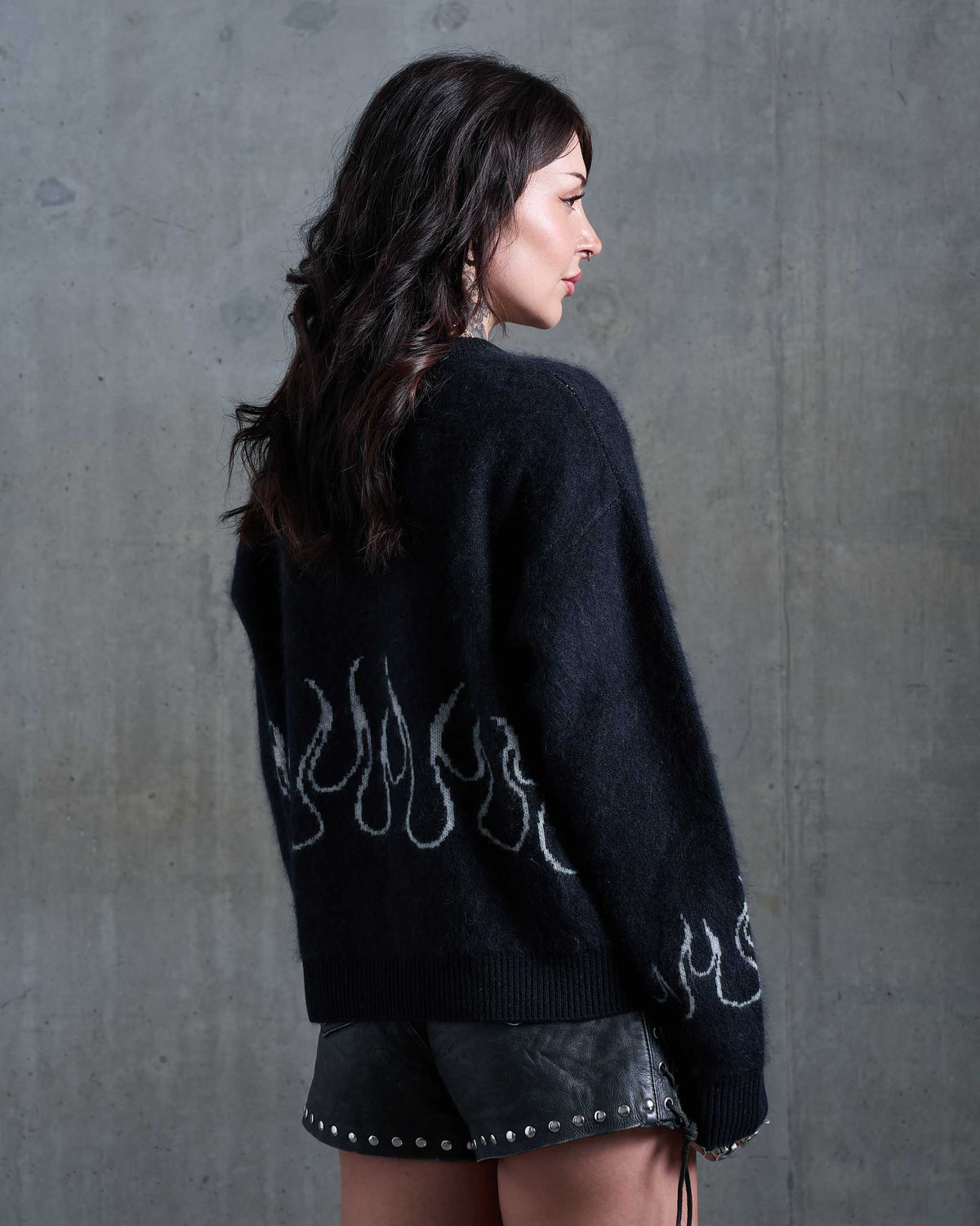 A woman wearing a black fuzzy knit sweater with white flame patterns on the back and sleeves, paired with black studded leather shorts, standing against a gritty concrete wall.