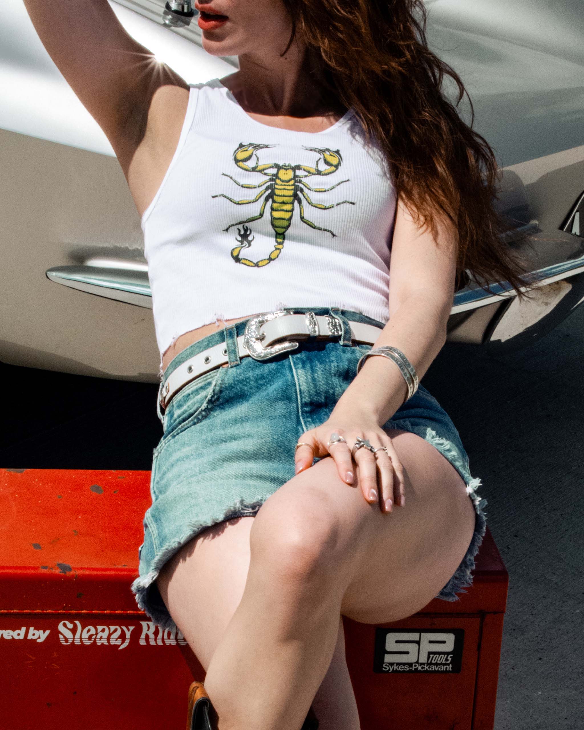 A model wearing a Sleazy Rider white tank top with a green scorpion design, denim shorts and a western belt, sitting by a classic car.