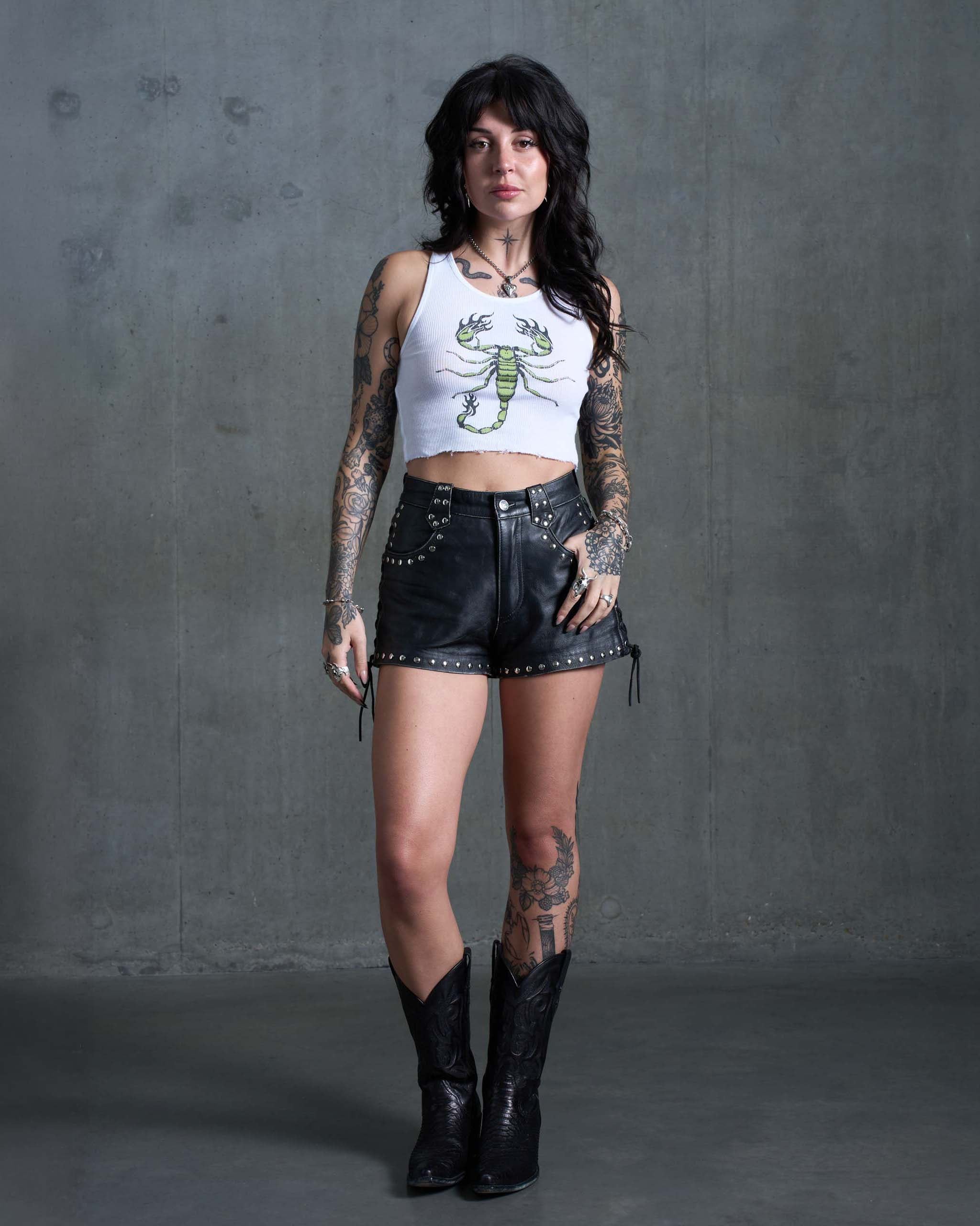 A tattooed woman wearing a green scorpion tank top, studded leather shorts and black cowboy boots.