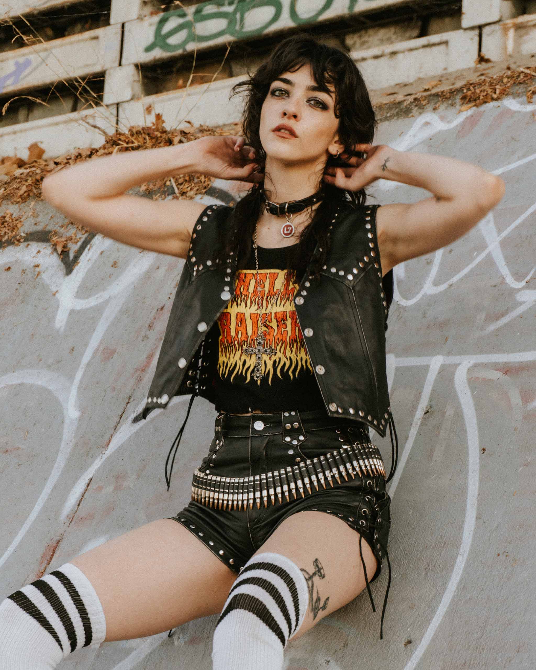Alt person wearing a gothic studded black leather outfit with a "Hell Raiser" graphic tank top, sitting against a graffiti-covered wall.