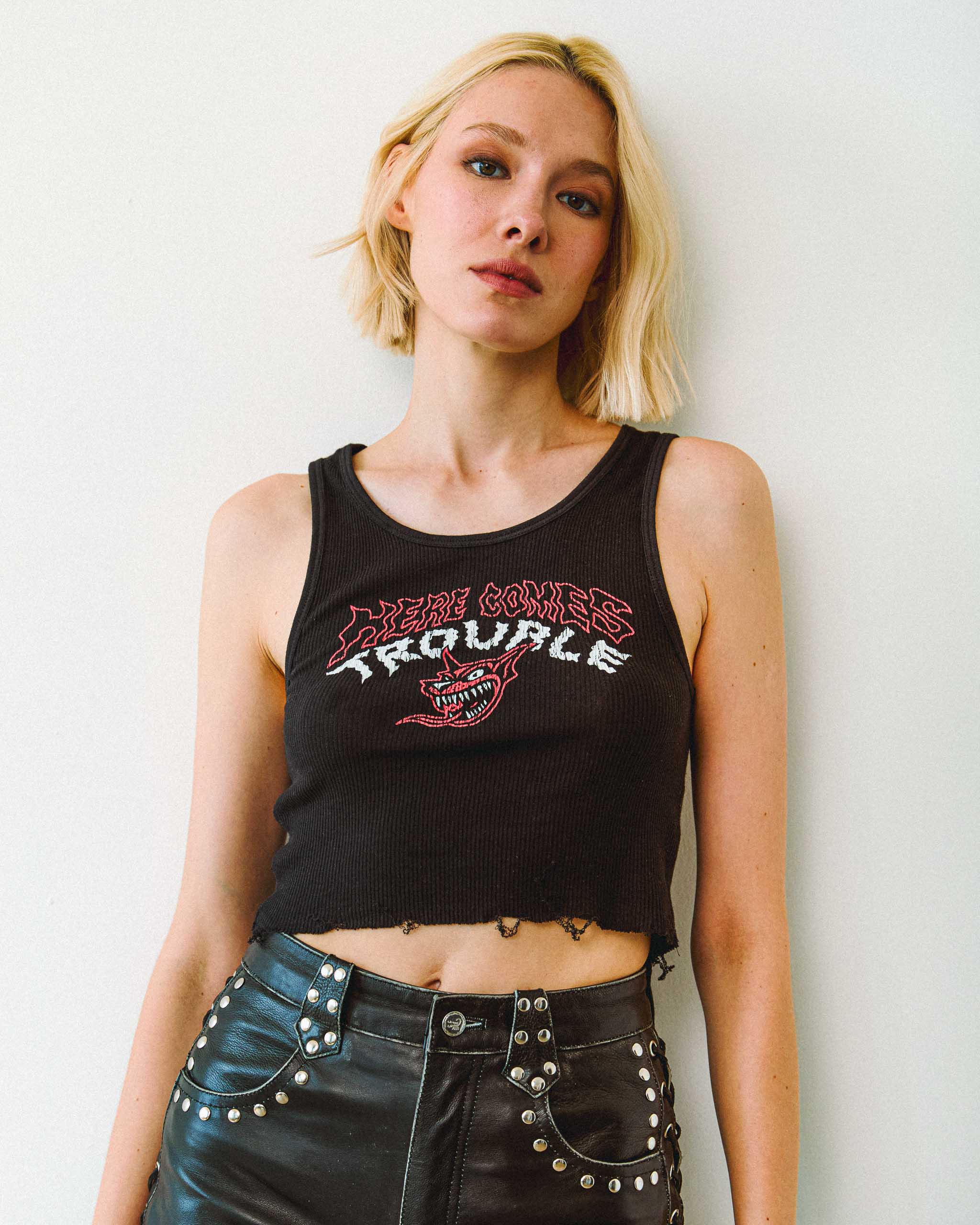 A young model wearing a black tank top with a vintage "Here Comes Trouble" print and studded leather shorts, on a plain background.