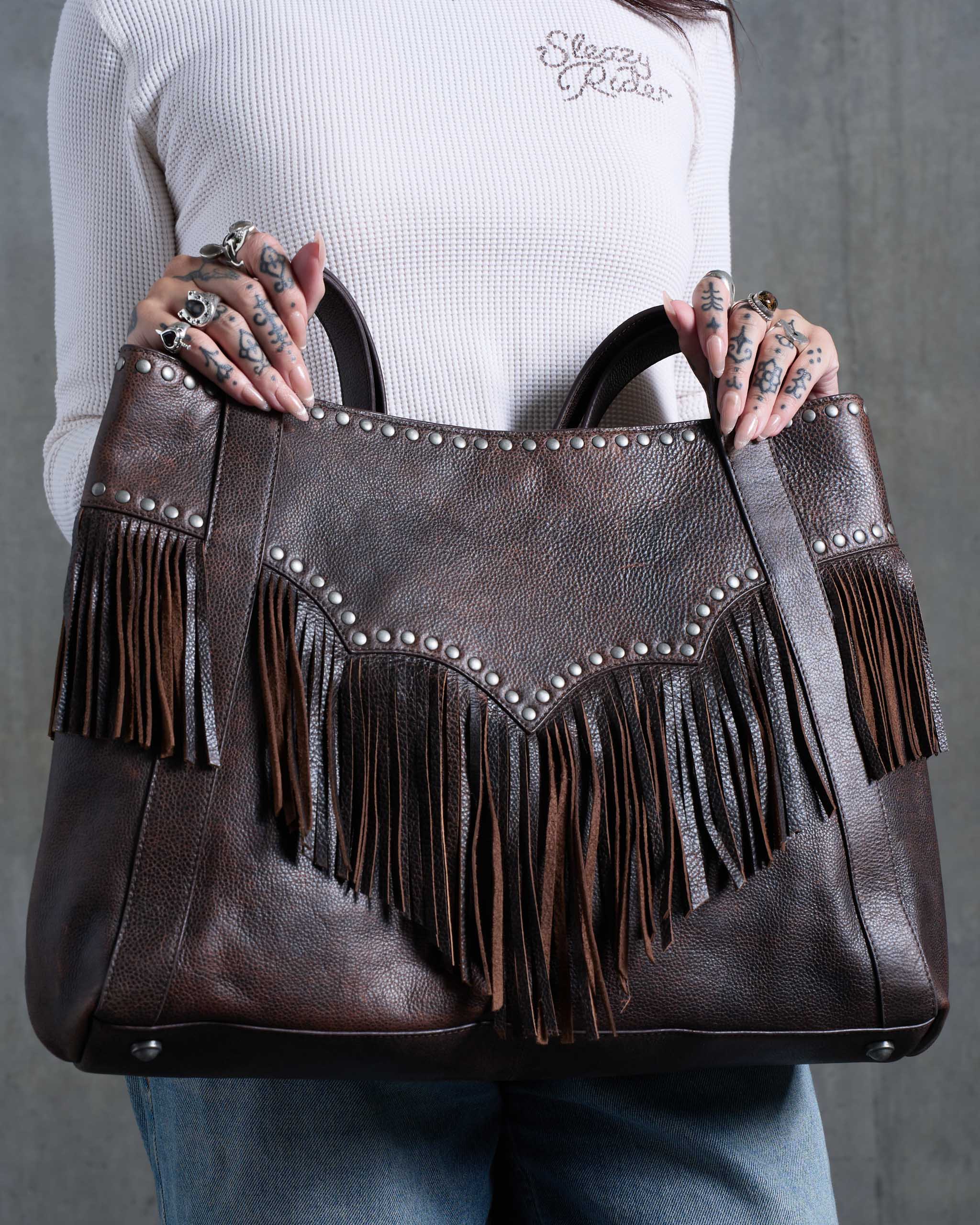A woman holding a large brown western style leather bag with studs, fringe and western yoke detailing.