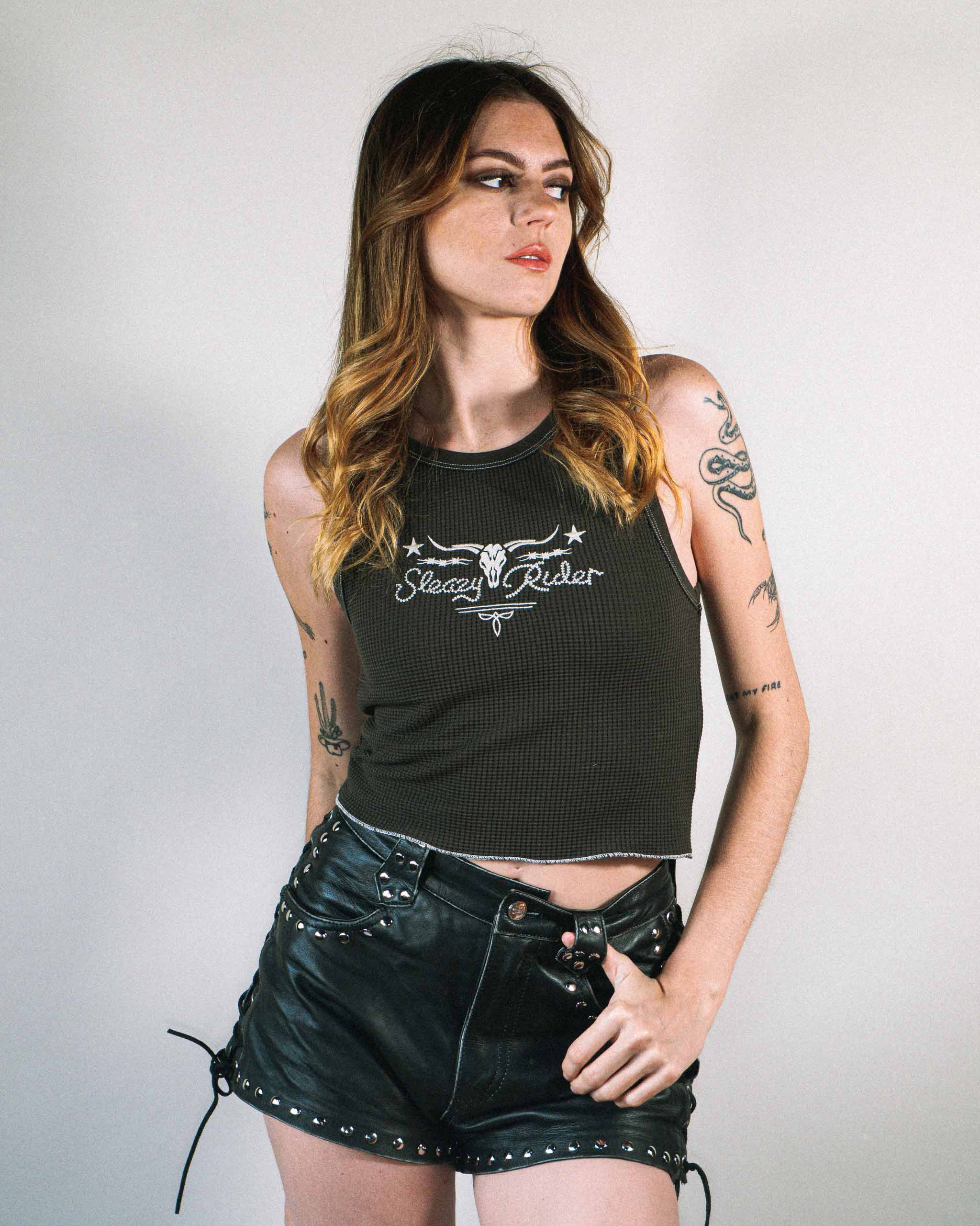 An alternative female model wearing a dark western tank top with cow skull embroidery and studded leather shorts.