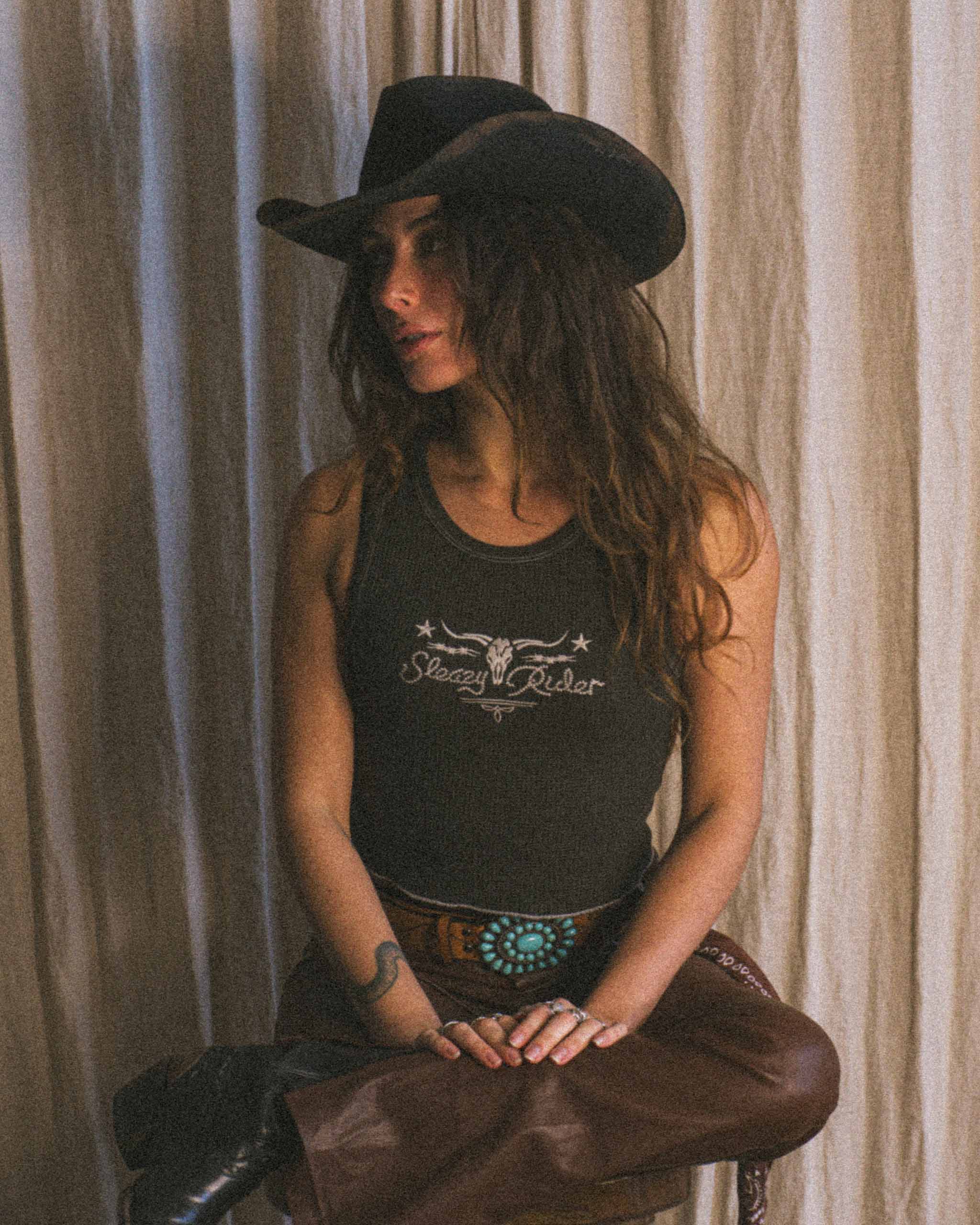A cowgirl wearing a cowboy hat and a western tank top, sitting on a bar stool.