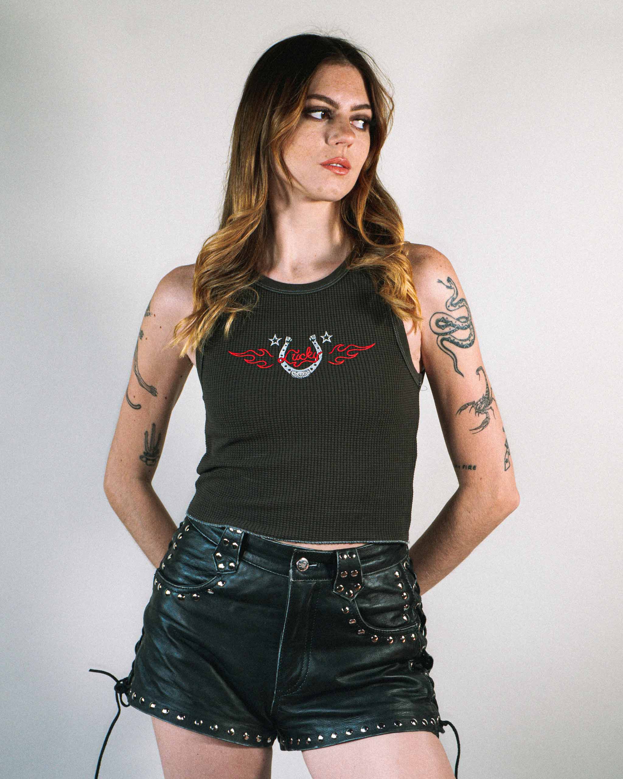 Alt model wearing a dark western tank top with a flaming horseshoe embroidery and studded leather shorts.