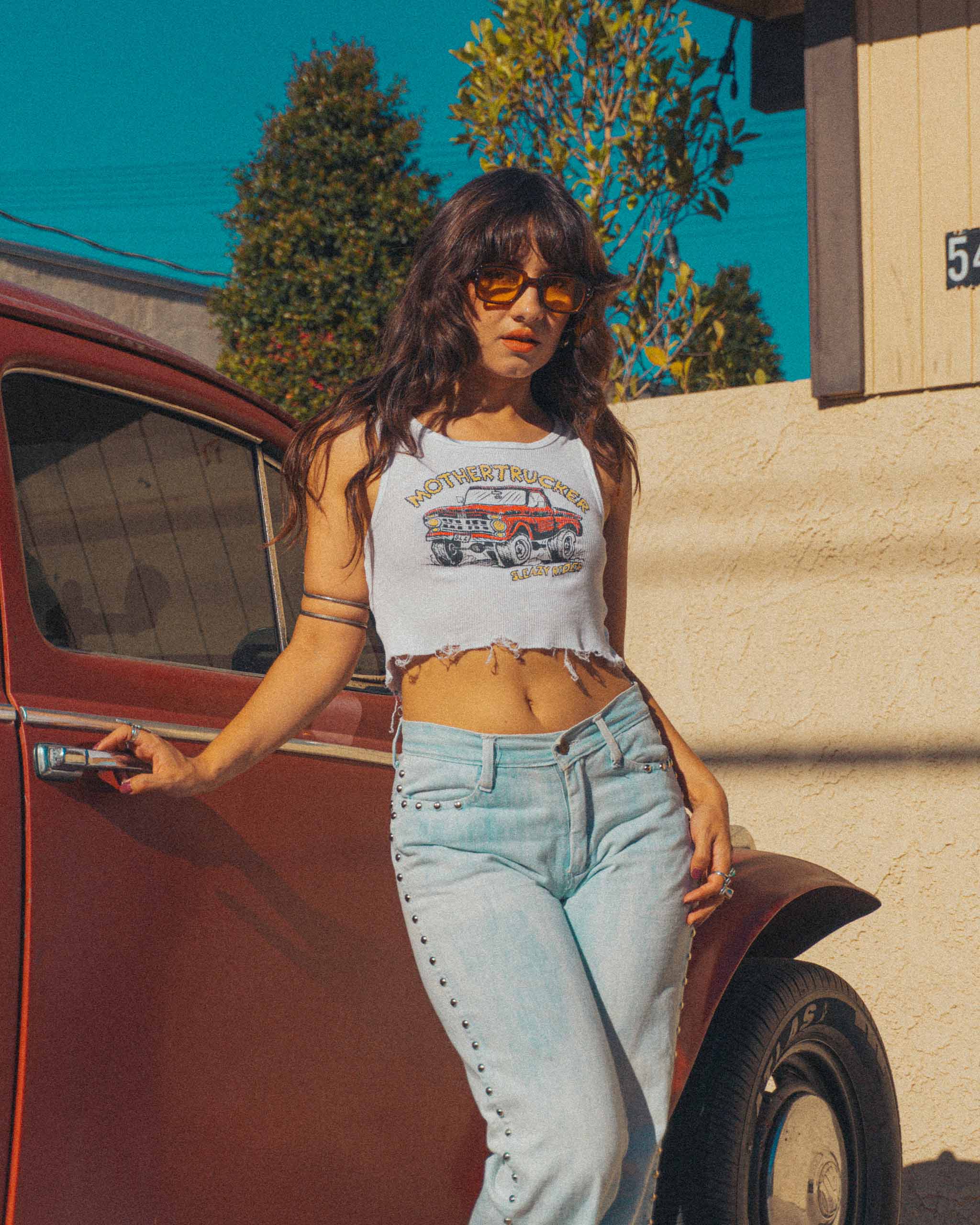 Woman standing next to a vintage car wearing a "Mothertrucker" graphic tank crop top and studded blue jeans.