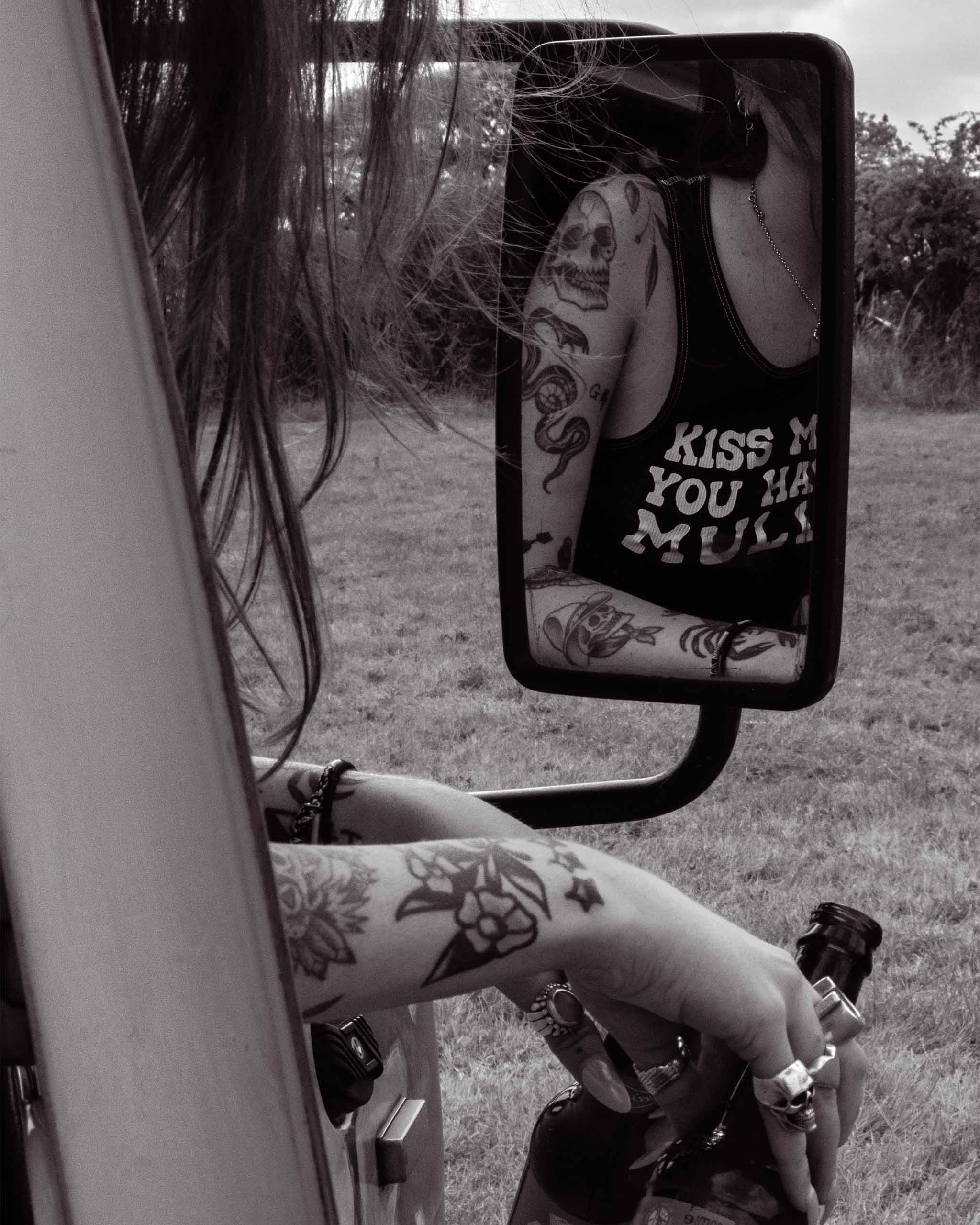 Person with tattoos sitting in a vehicle, reflected in a mirror wearing a tank top that says "Kiss Me if you Have a Mullet".