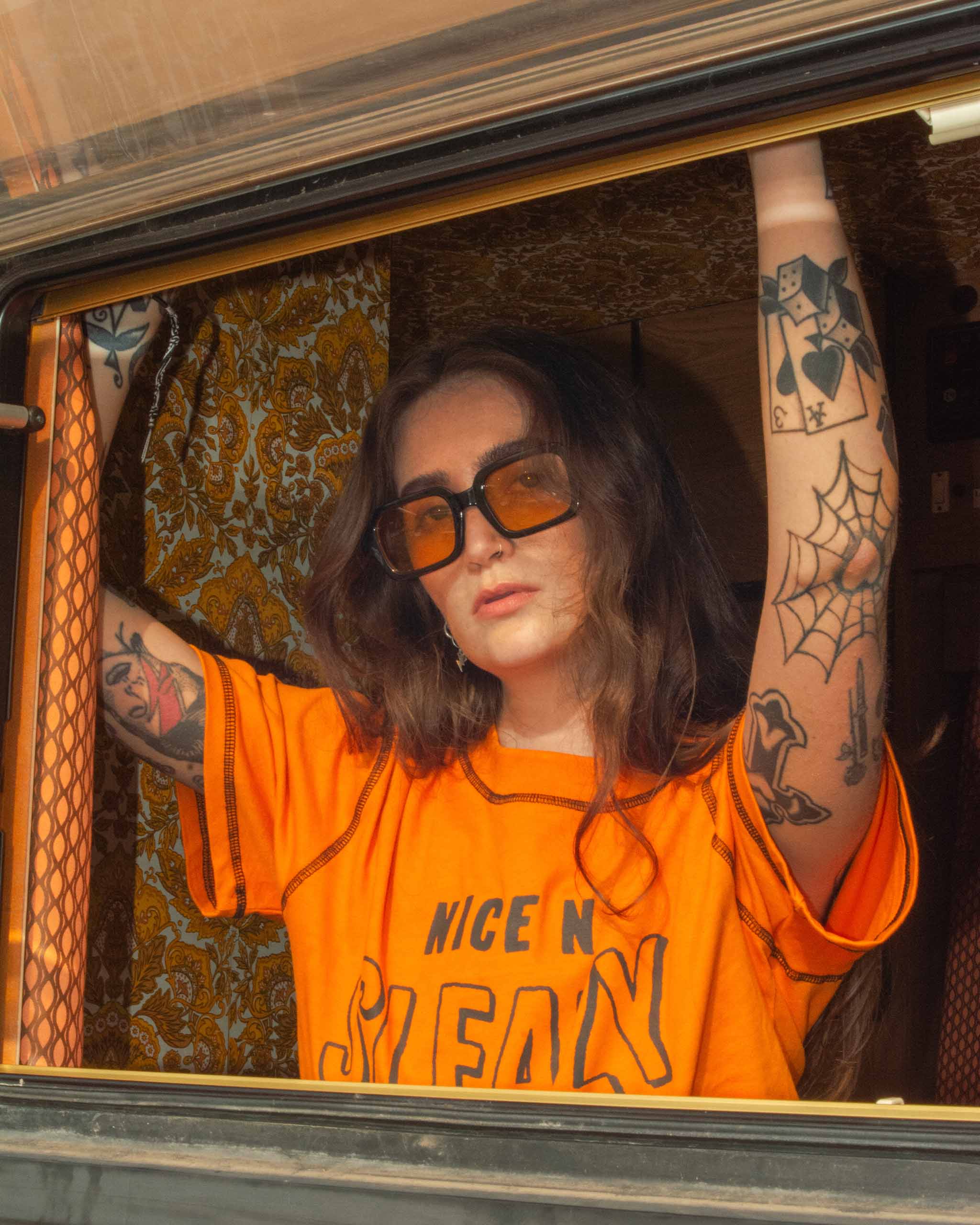A woman in a 70s campervan wearing a bright orange t-shirt with "Nice n' Sleazy" text in a chunky black vintage font and 70s orange sunglasses.