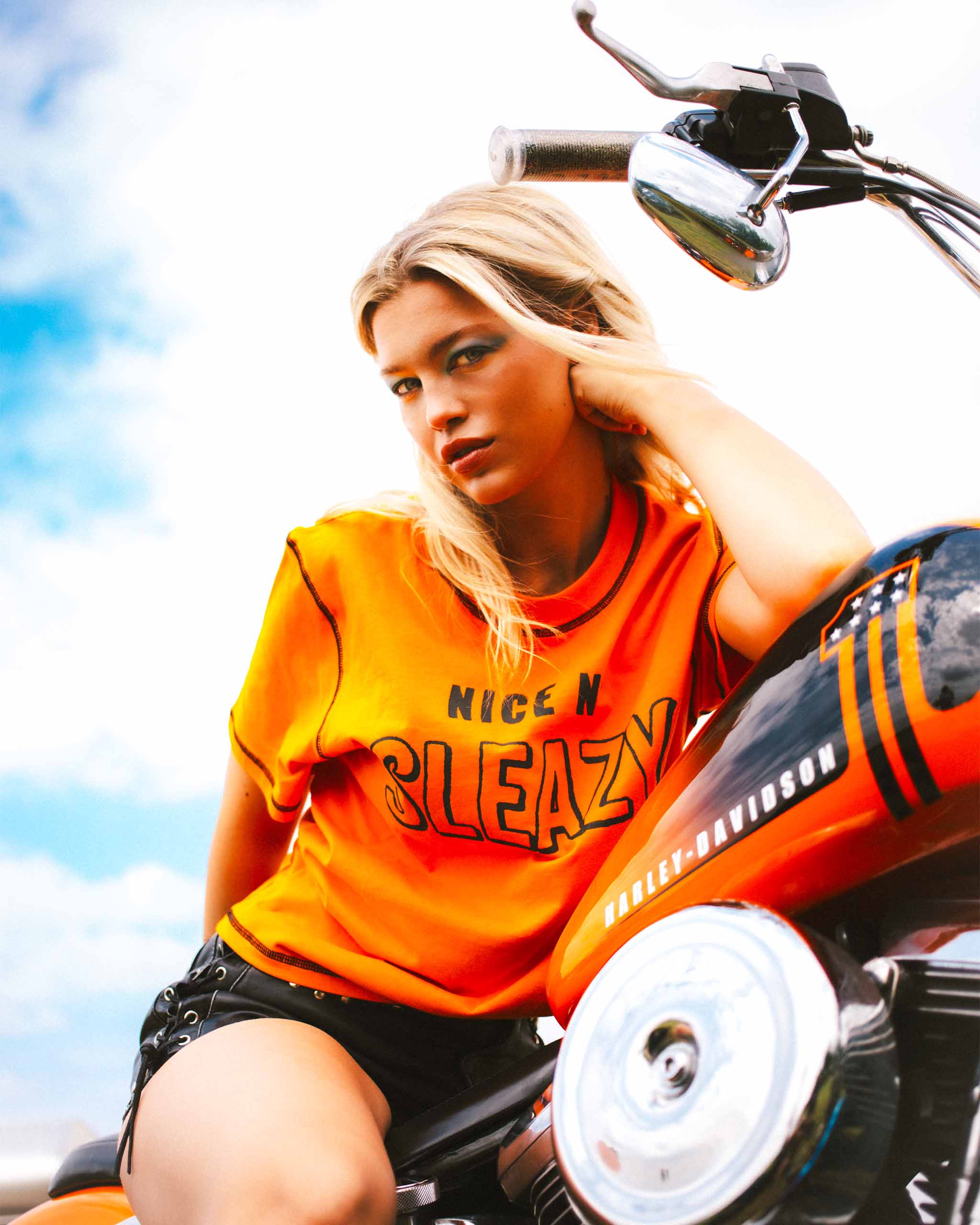 A model on an orange motorcycle wearing a bright orange "Nice n' Sleazy" slogan t-shirt with contrast stitching.