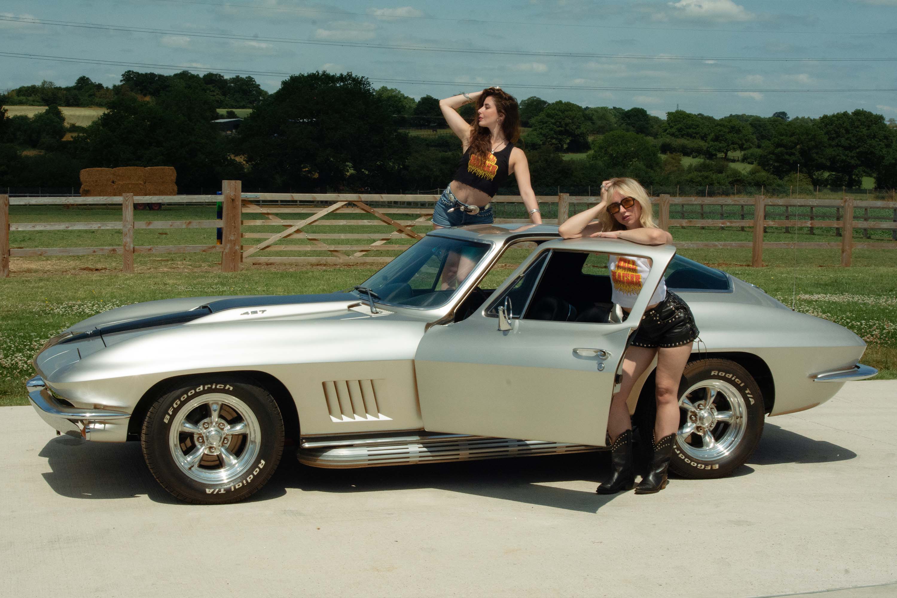 Two women wearing Sleazy Rider 'Hell Raiser' posing next to a classic silver Corvette.