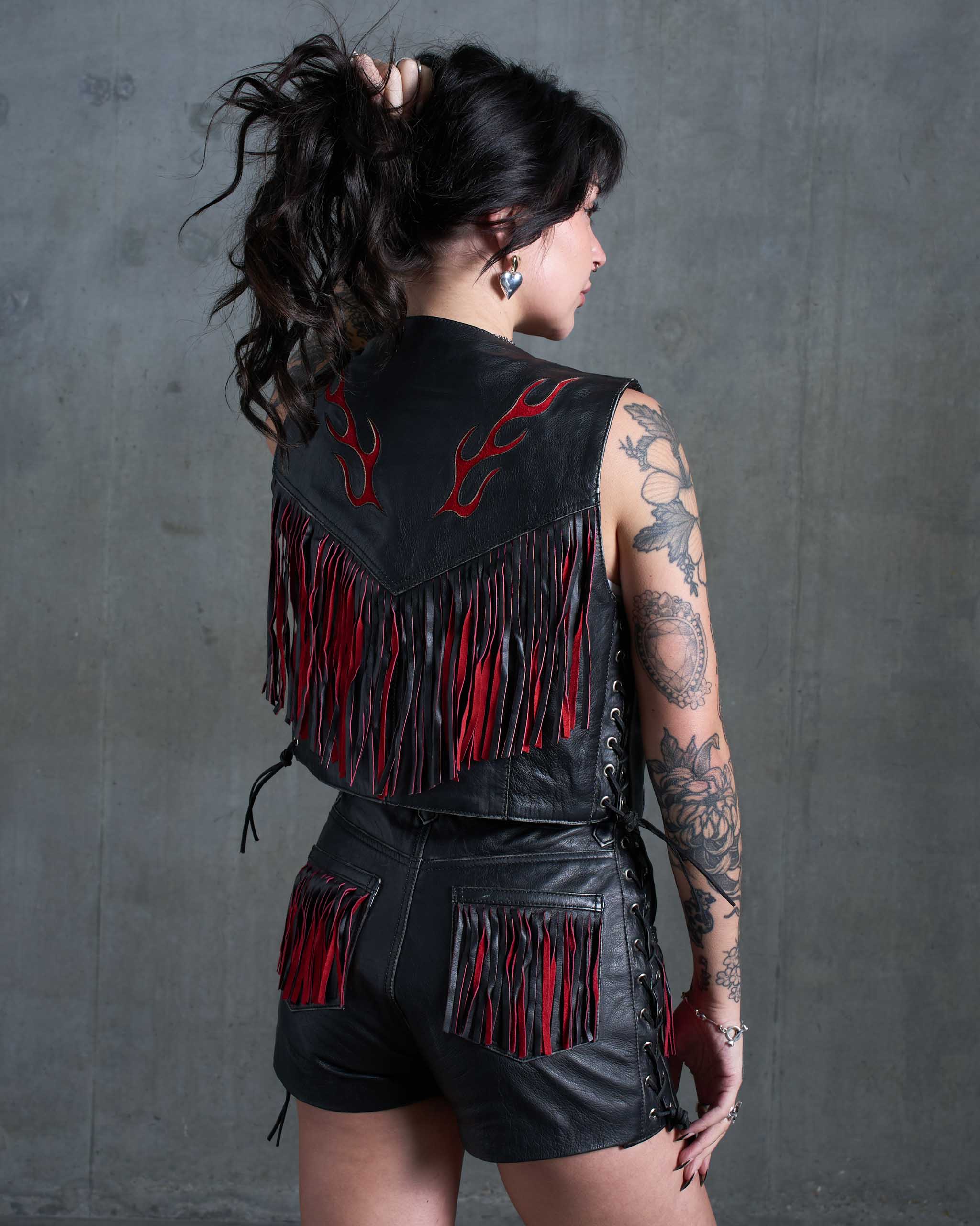 A tattooed model wearing a biker leather vest and shorts set with red flames details.