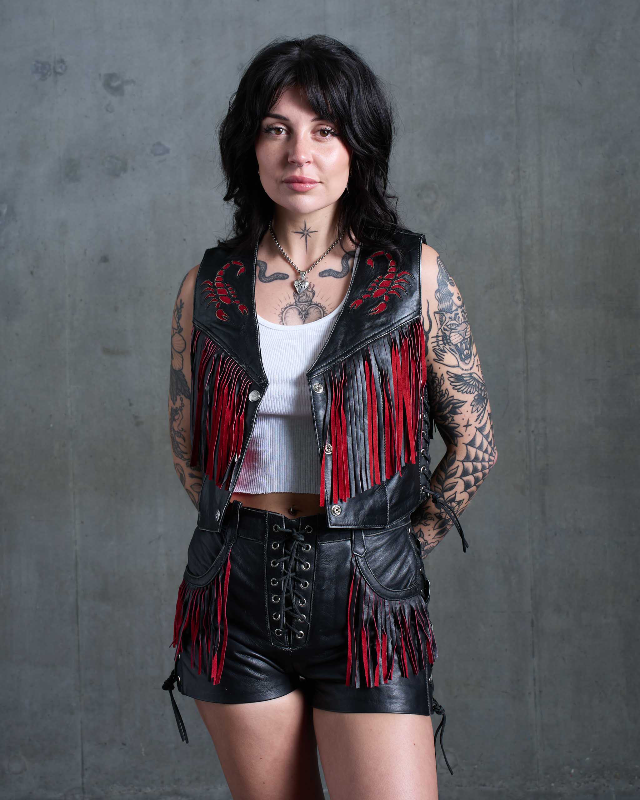 Woman wearing a black leather vest with red fringe and red suede scorpion details, standing against a concrete background.