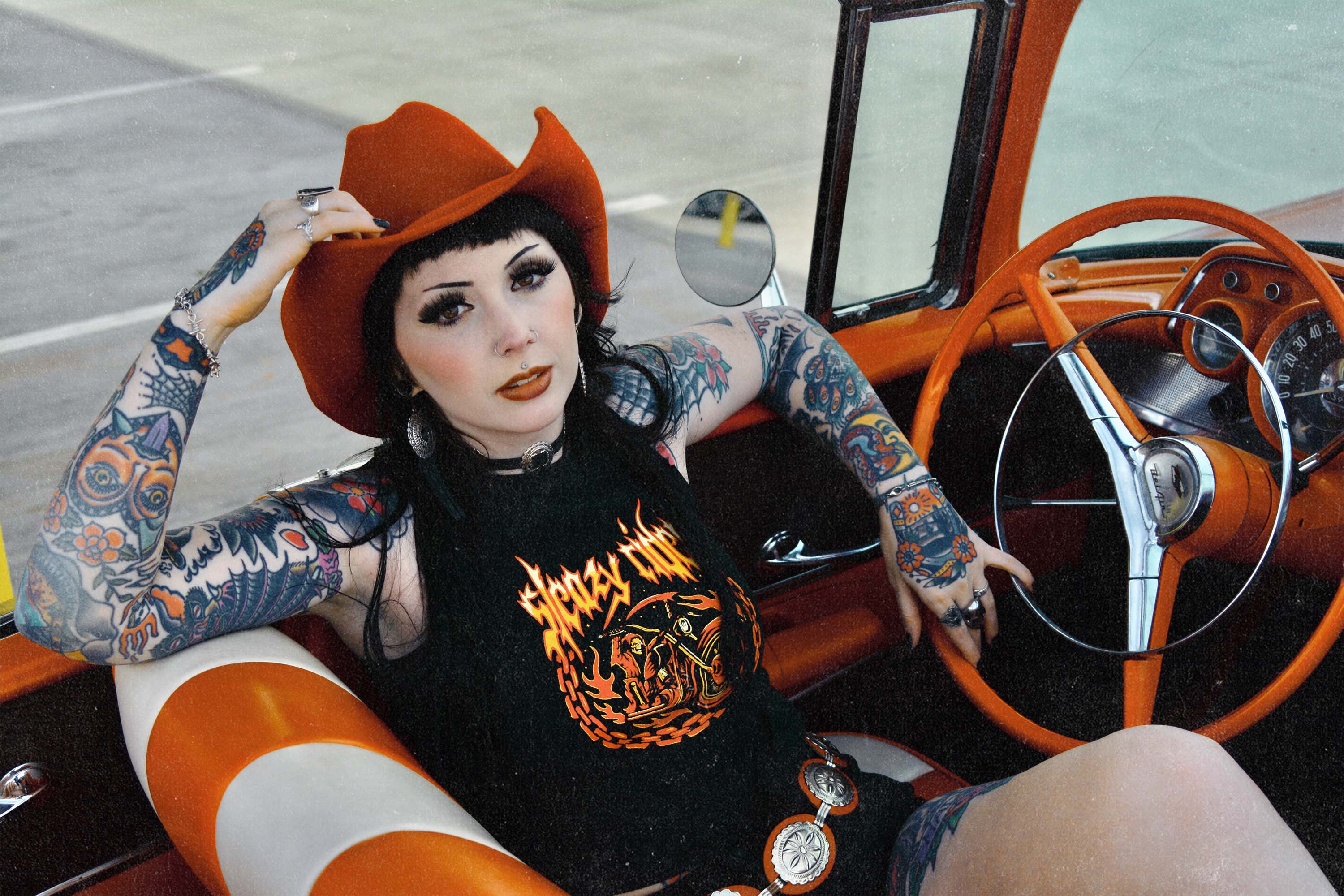 A tattooed alternative model wearing a Sleazy Rider tank top, in a vintage car