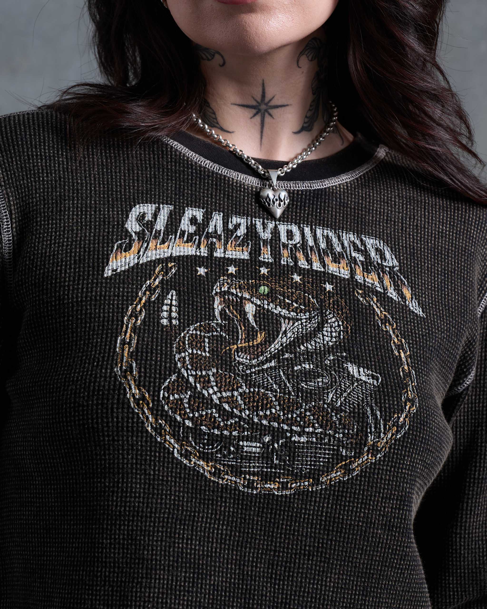 A close-up of a tattooed woman wearing an acid wash waffle knit sweater with a rattlesnake print on the chest, and contrast stitching.