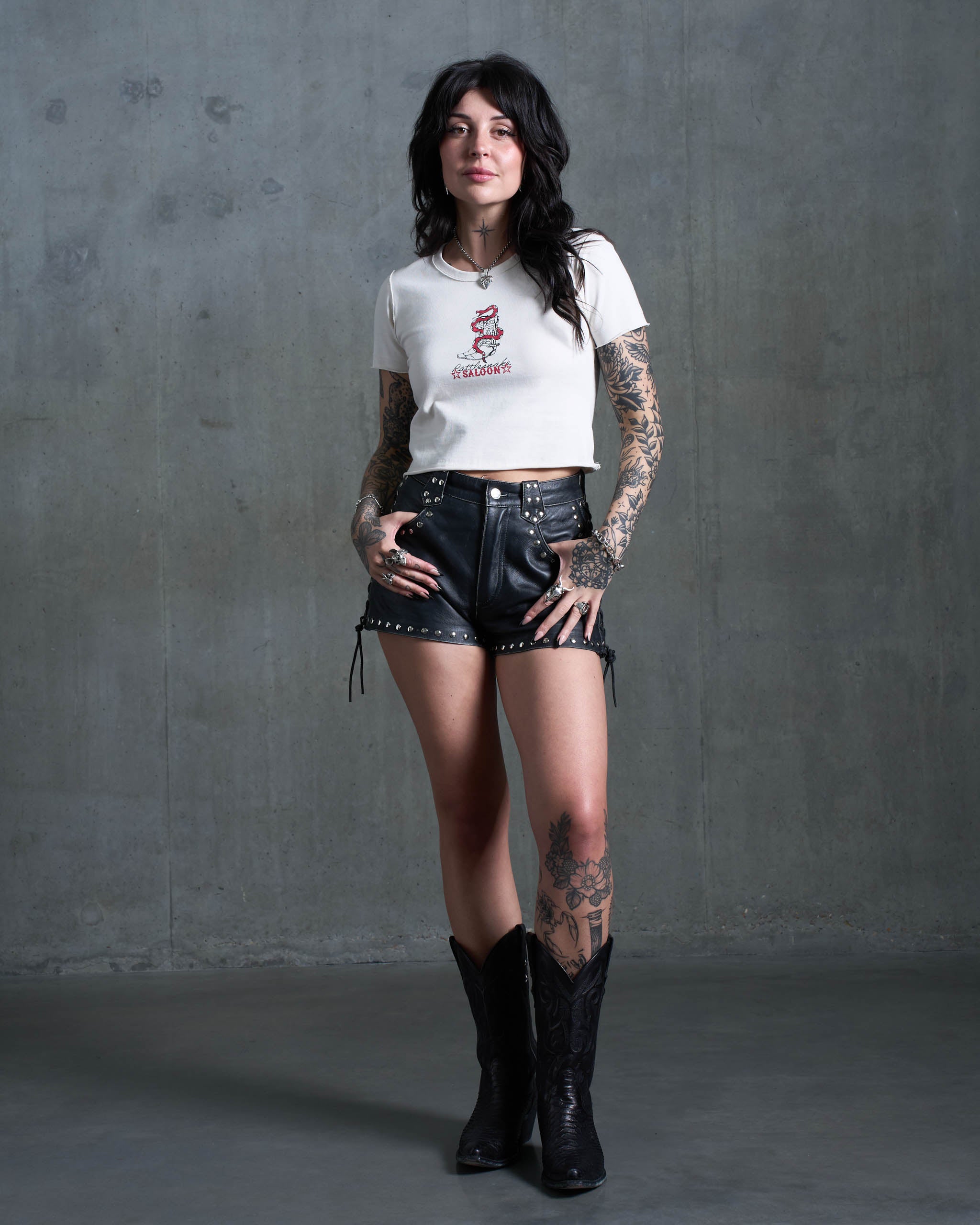 An alt western woman wearing a rattlesnake saloon baby tee, studded leather shorts and cowboys boots.
