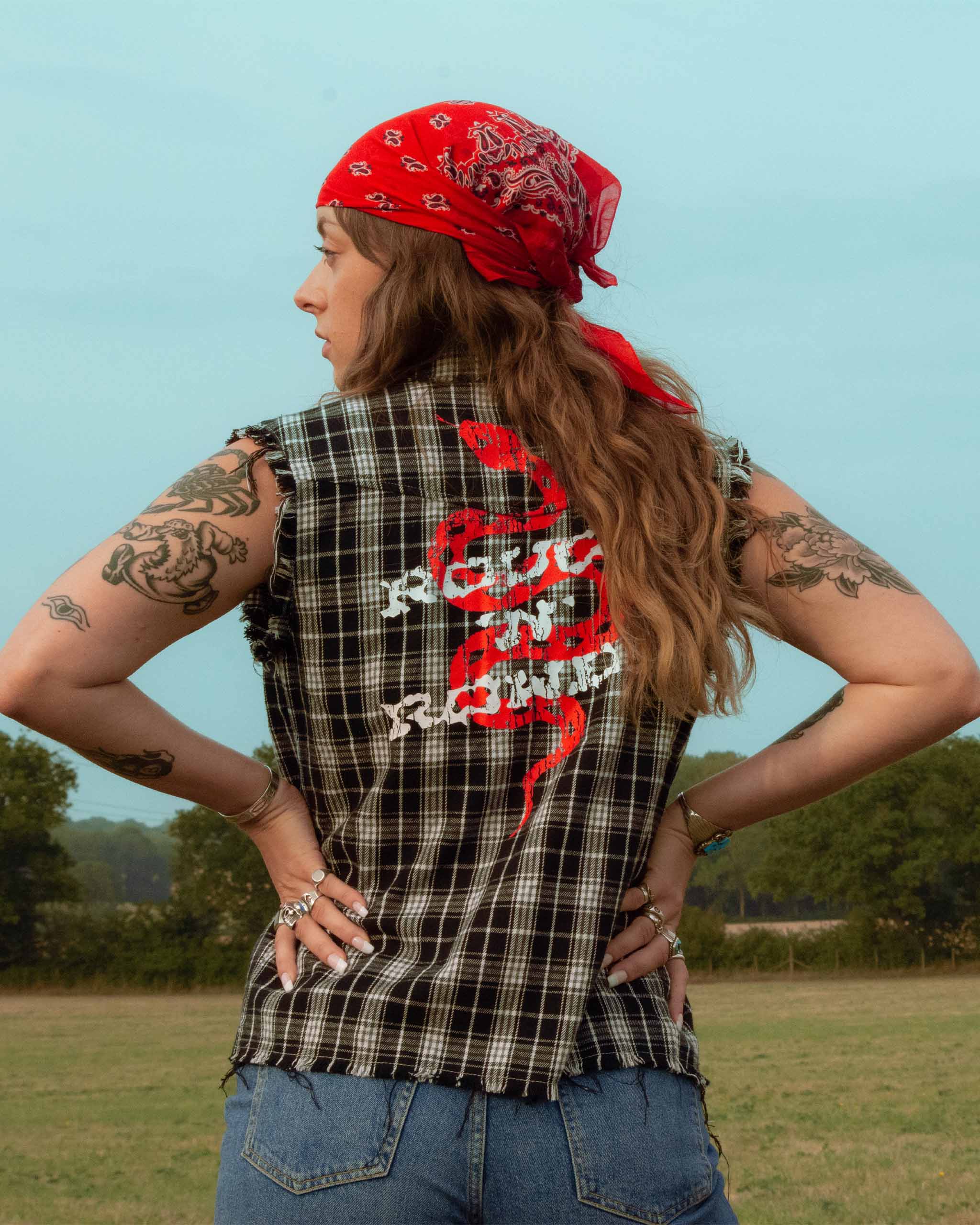 Person wearing a plaid black and white sleeveless shirt with snakeprint graphic and a red bandana outdoors.