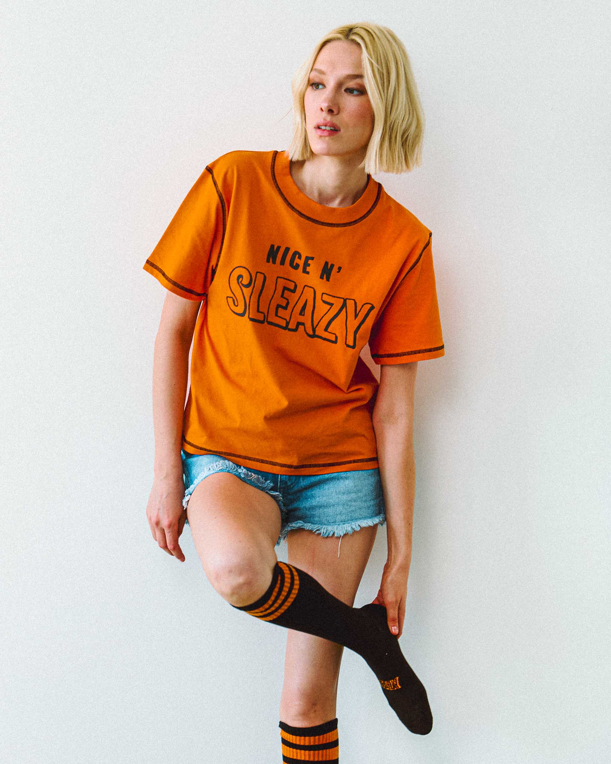 Woman wearing an orange "Nice N' Sleazy" t-shirt and black knee-high Sleazy Rider socks with orange stripes on a white background.