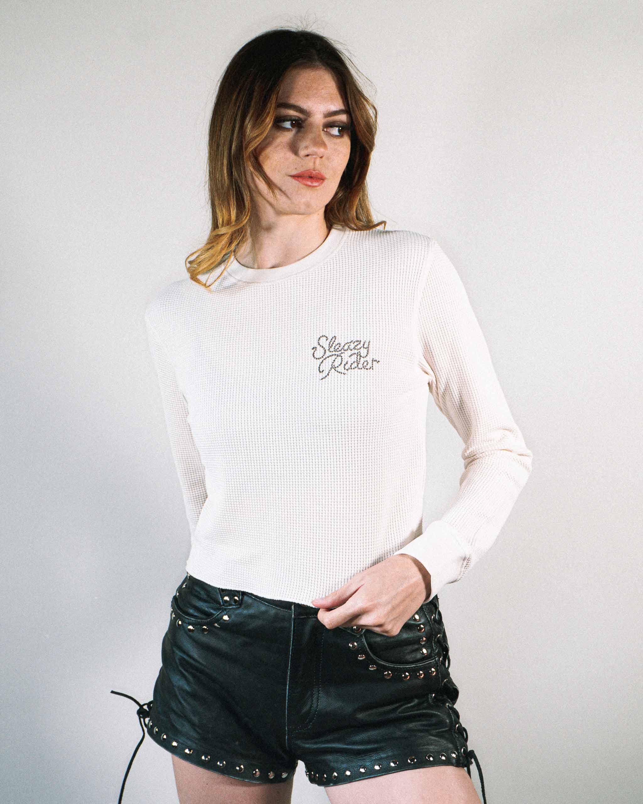 An alt model wearing an off white waffle knit top with Sleazy Rider rope text and leather shorts.